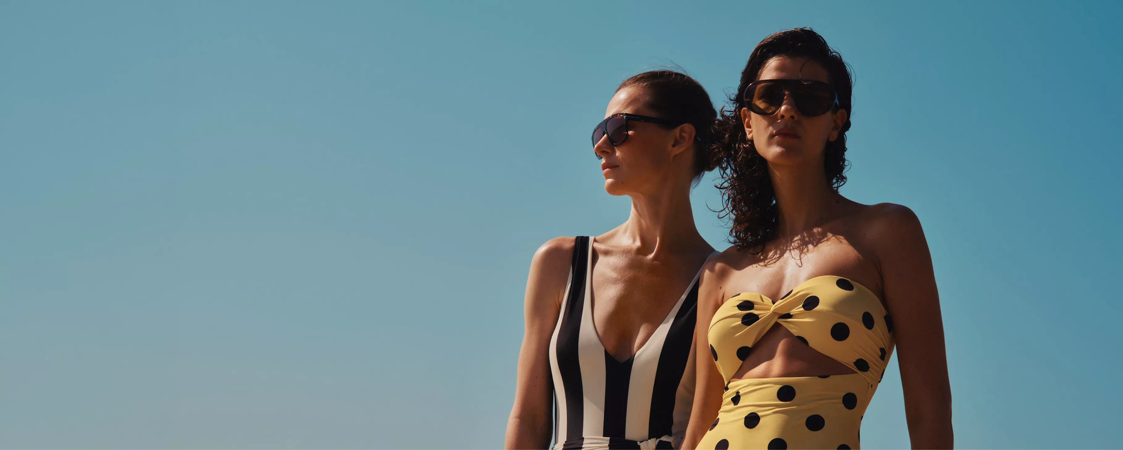Two women in stylish swimsuits and sunglasses posing under clear blue sky, summer beach fashion editorial shot