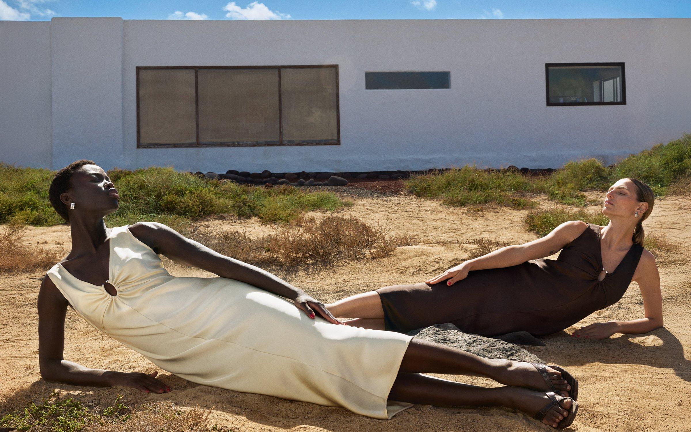 2 women dressed in formal dresses lying on a rocky surface with a white building and bright blue sky behind them