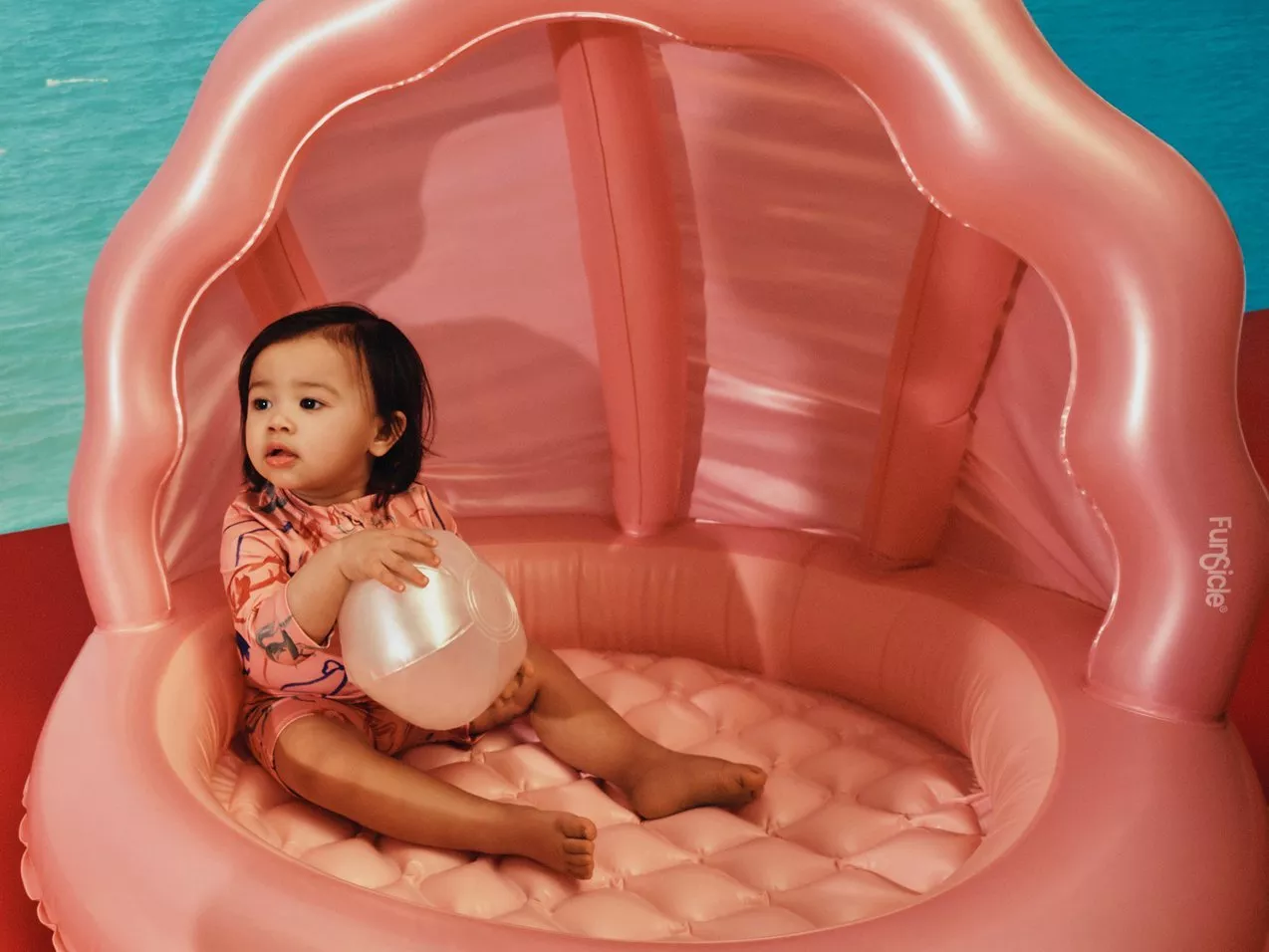 Toddler sitting in a pink inflatable pool with canopy, holding a beach ball beside turquoise water background