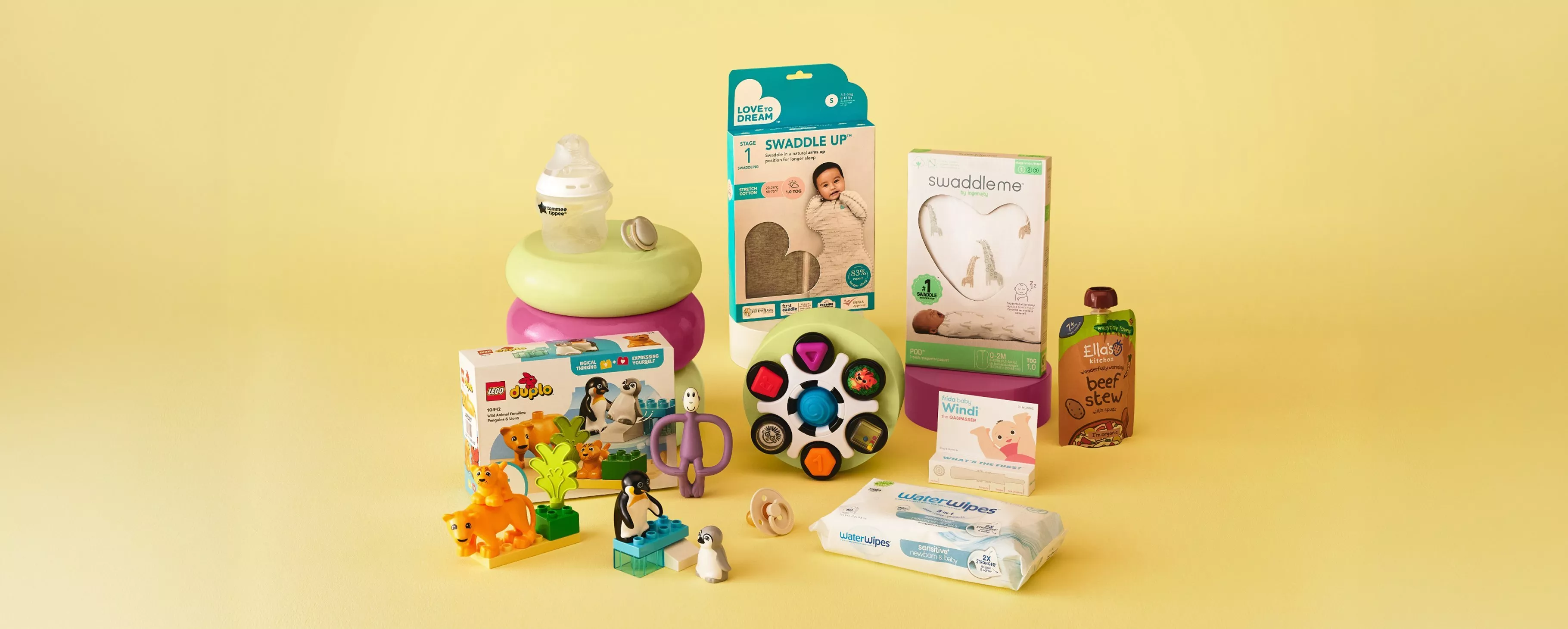 Baby essentials assortment with swaddle, nappies, wipes, toys, bottle and toddler food pouches on yellow background