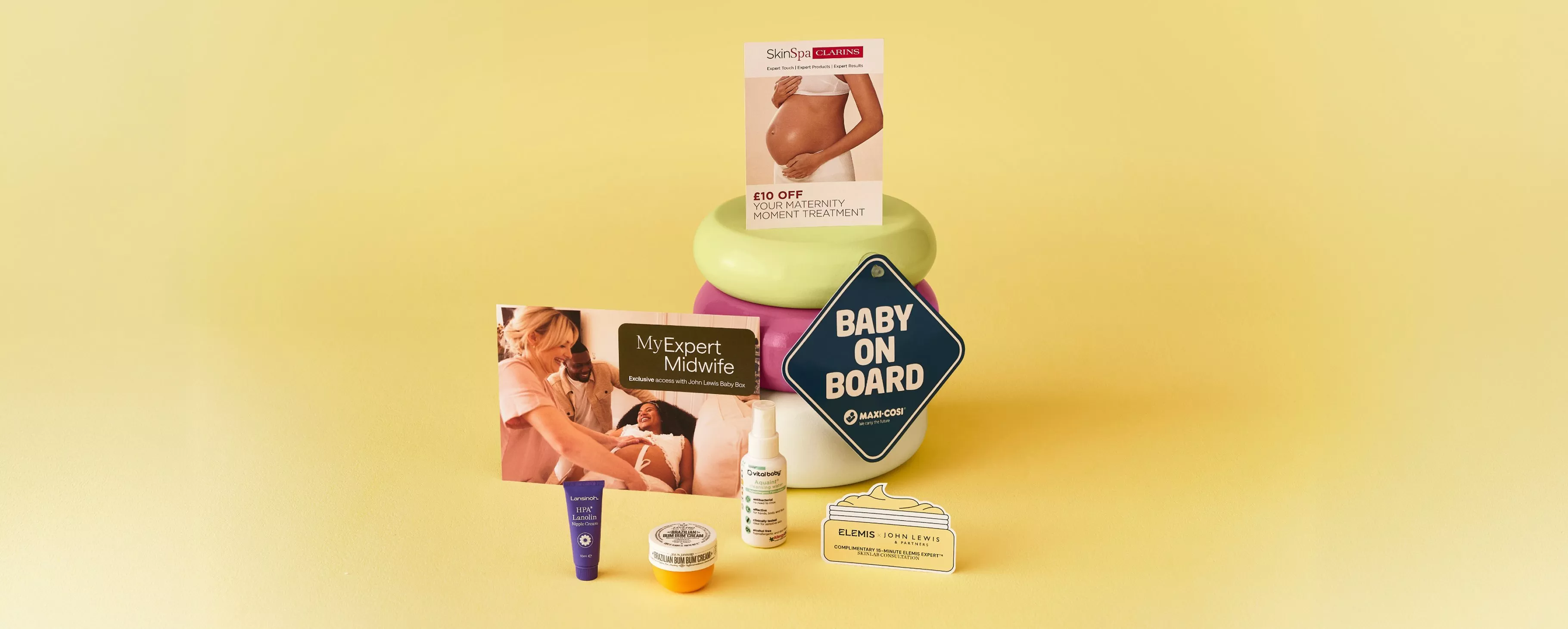 Pregnancy care gift set with skincare samples, midwife booklet and “Baby on Board” sign on yellow background