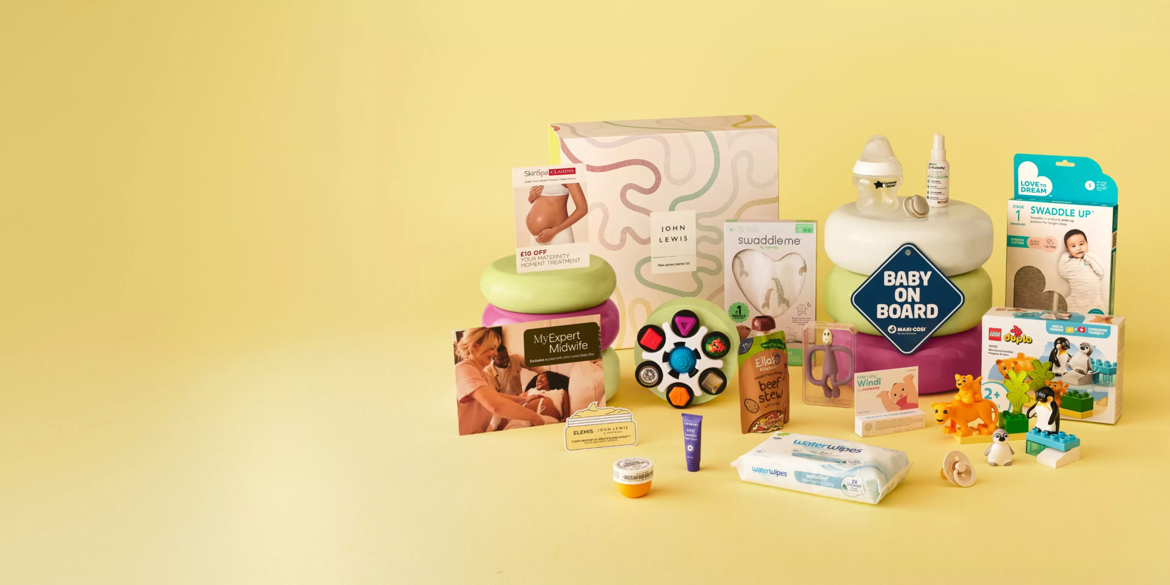 New baby gift hamper with swaddle, baby on board sign, toys, wipes and parenting essentials on yellow background