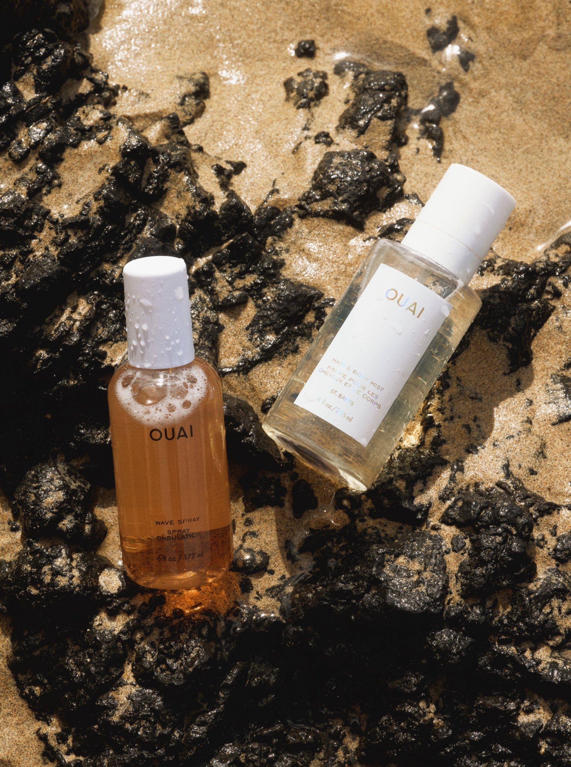 OUAI Wave Spray and OUAI Hair & Body Mist laid on a rock with some beach sand