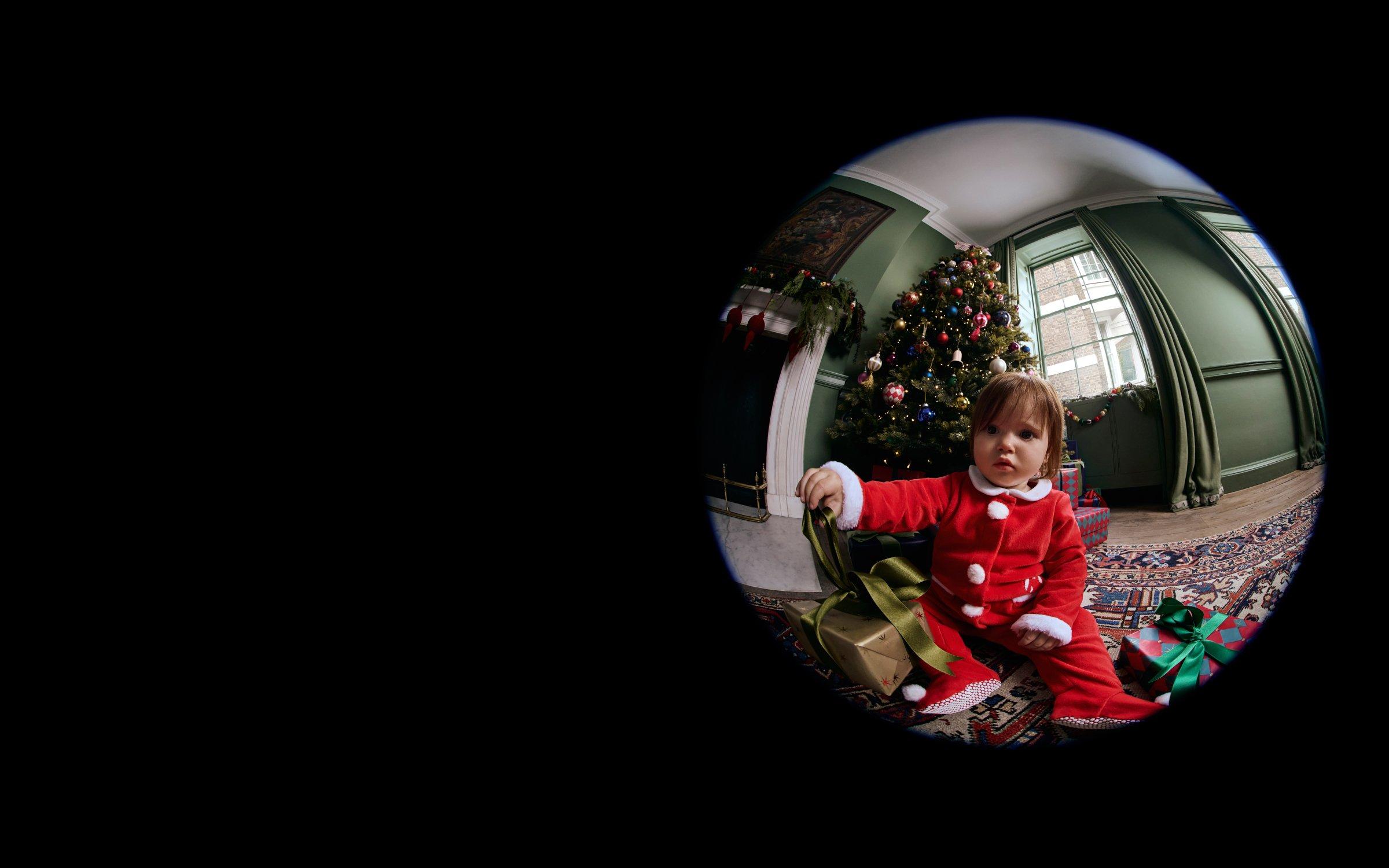 Fisheye lens image of a girl in a Santa Claus outfit sitting in front of a decorated Christmas tree.