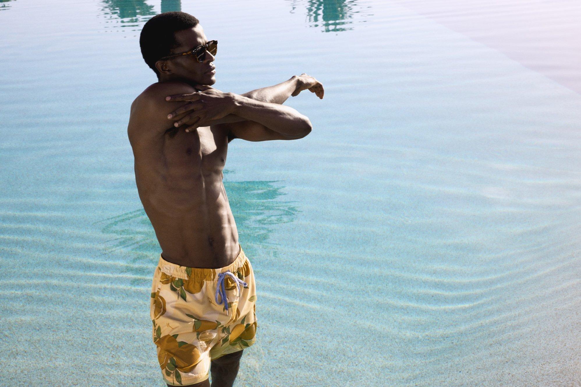 A male model wearing yellow swim shorts standing on a shallow beach