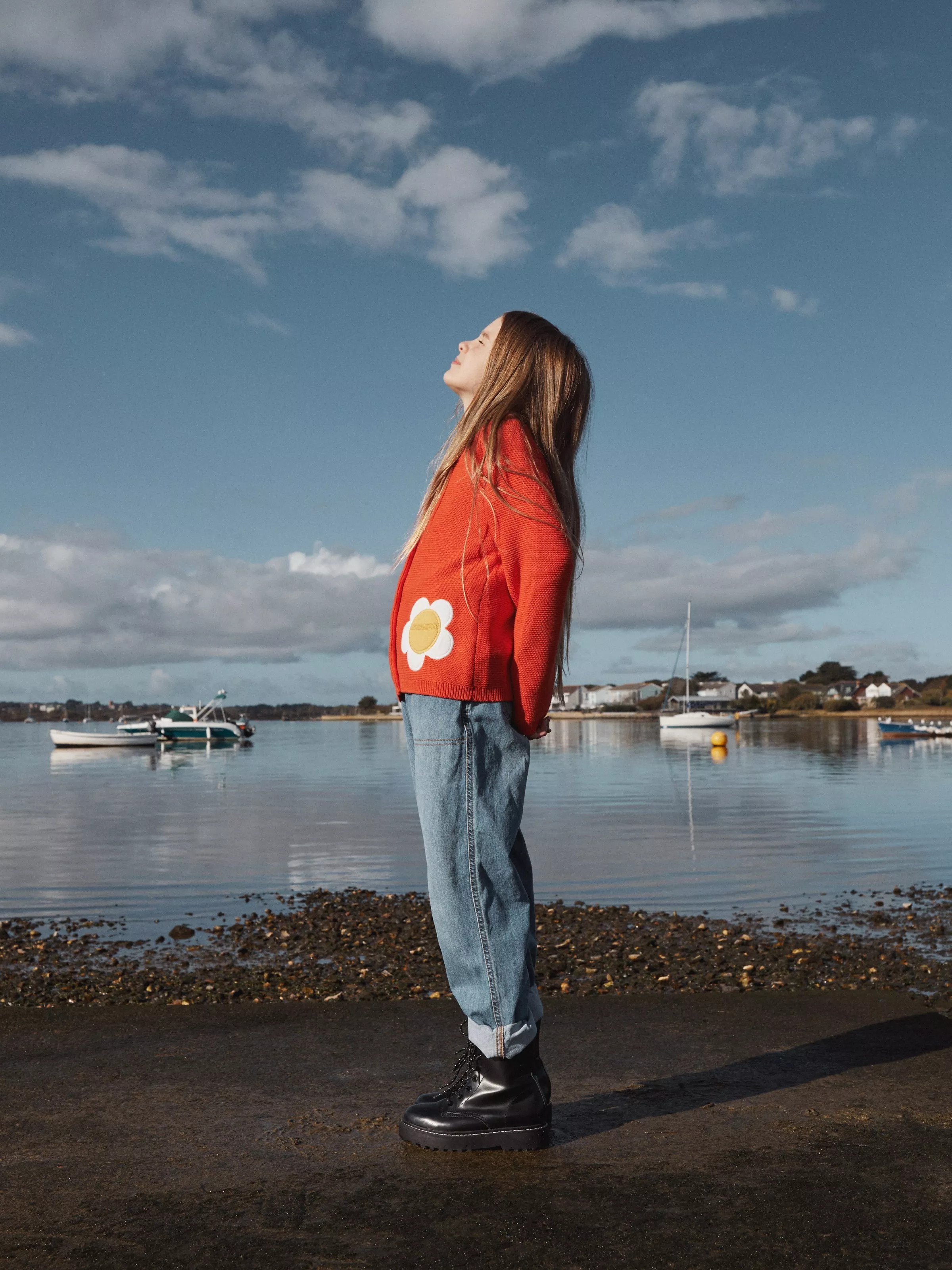 Girl in red jumper with daisy motif standing by calm harbour, boats on water under blue sky with clouds