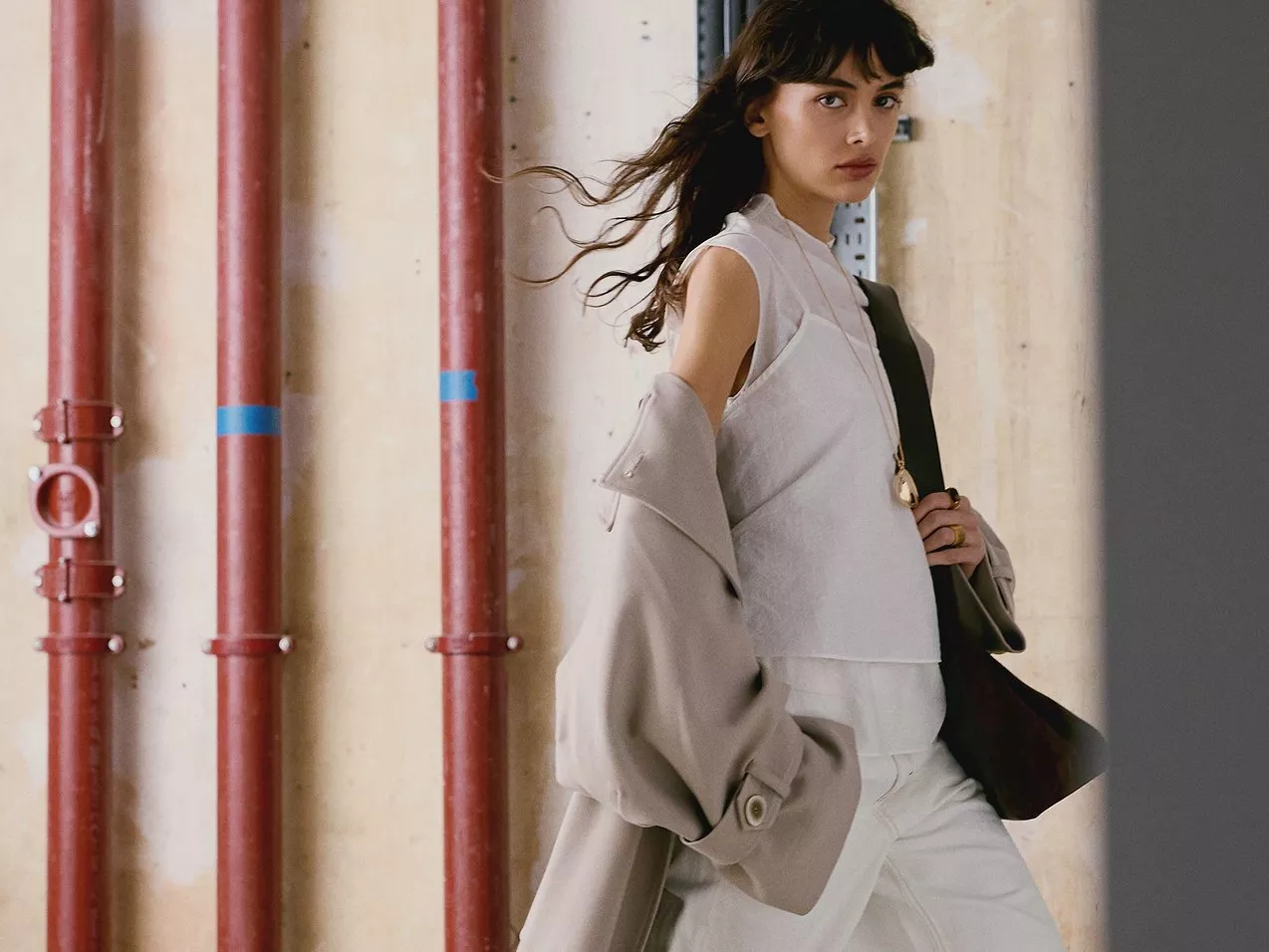 A woman in a beige coat and white outfit, standing by red pipes indoors.