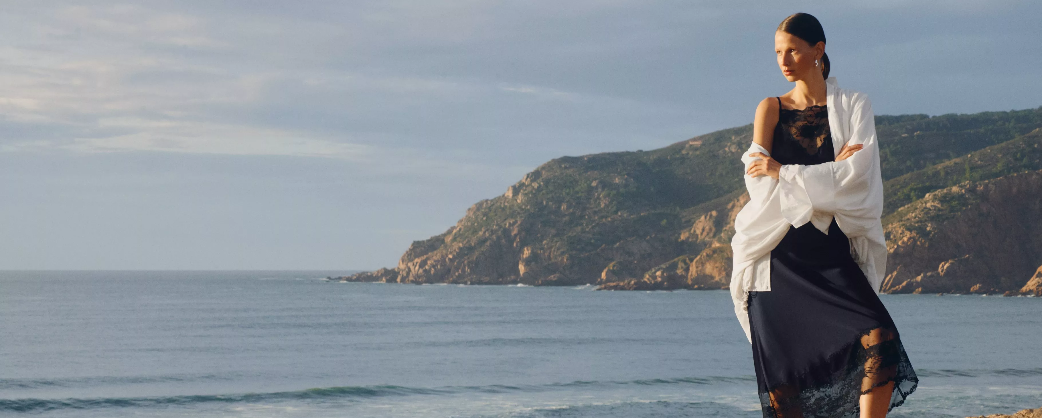 Woman in white jacket and navy blue pace dress, against a coastal backdrop.