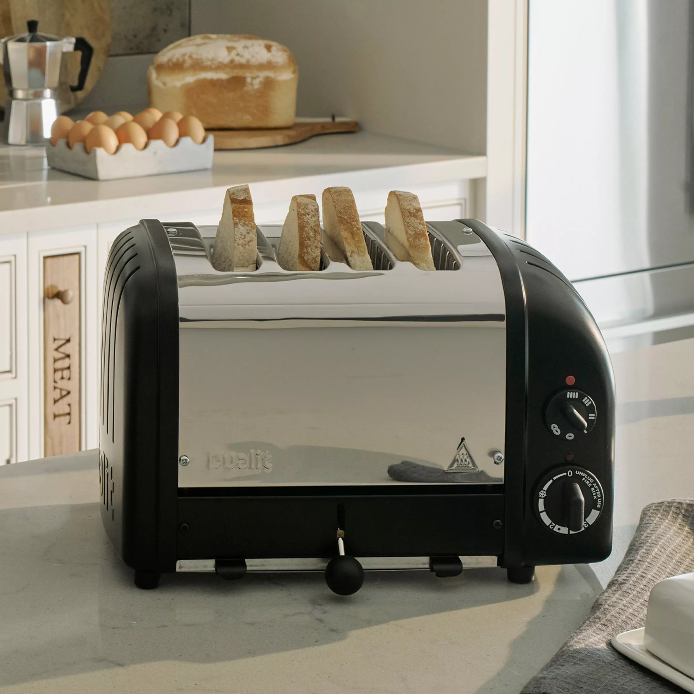 Four-slice stainless steel toaster with bread slices on a kitchen worktop, eggs and loaf in the background