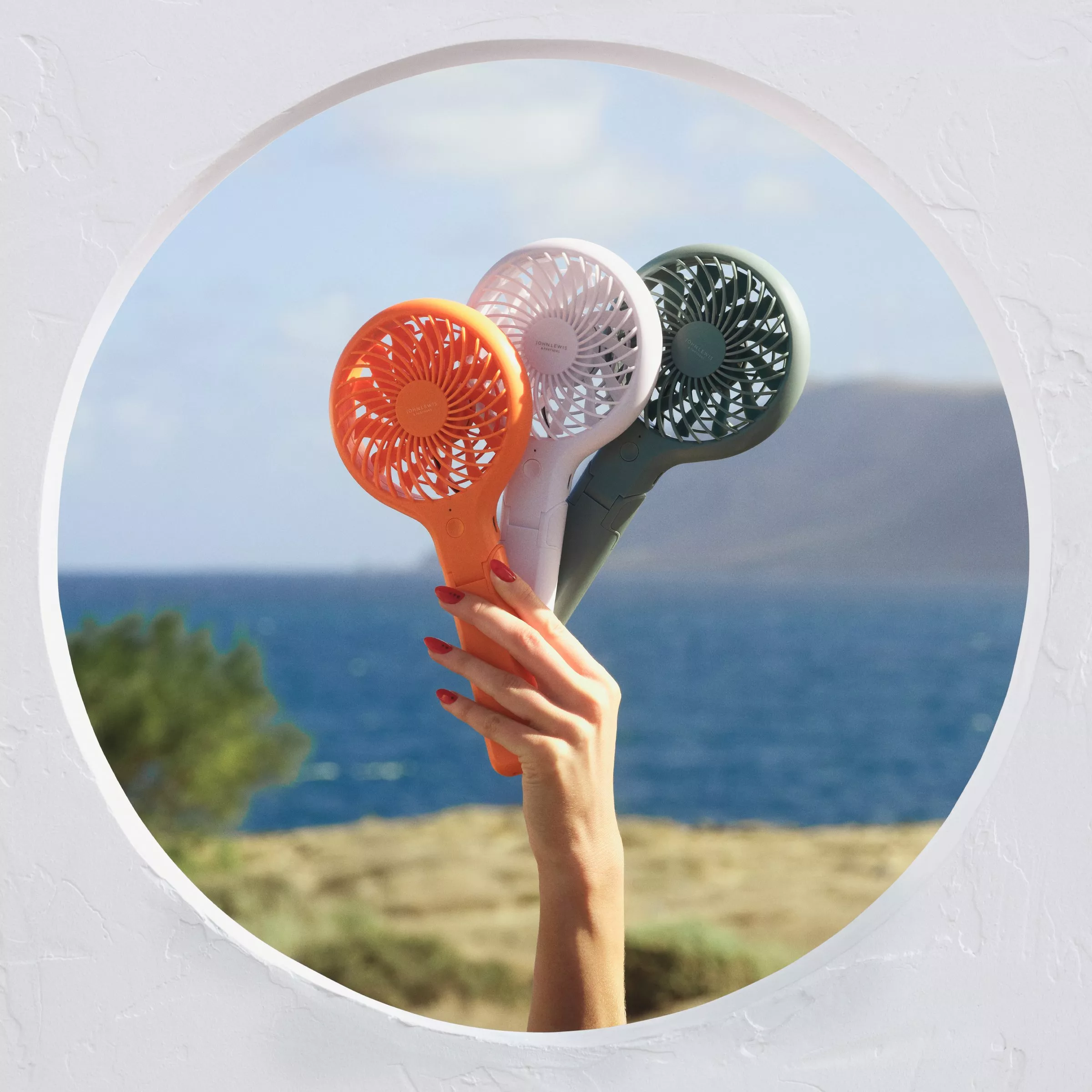 Hand holding three colourful portable mini fans against a seaside view, ideal for summer travel cooling on the go