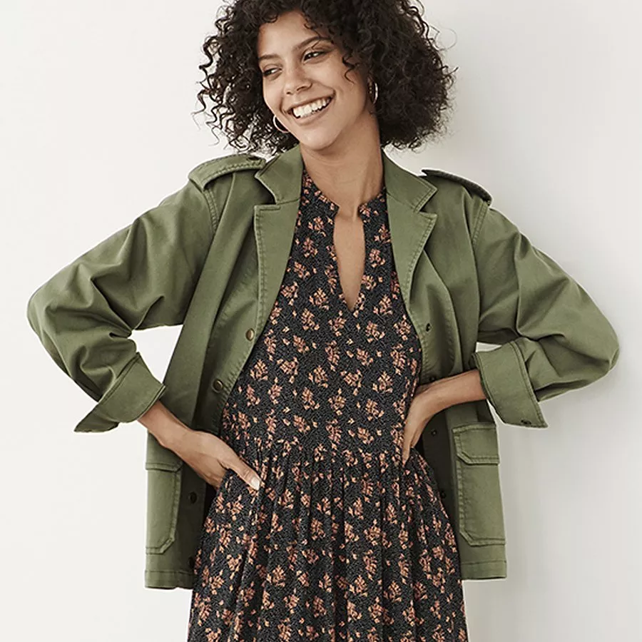 A person wears a green jacket over a floral dress, hands on hips, showcasing a casual yet stylish outfit.