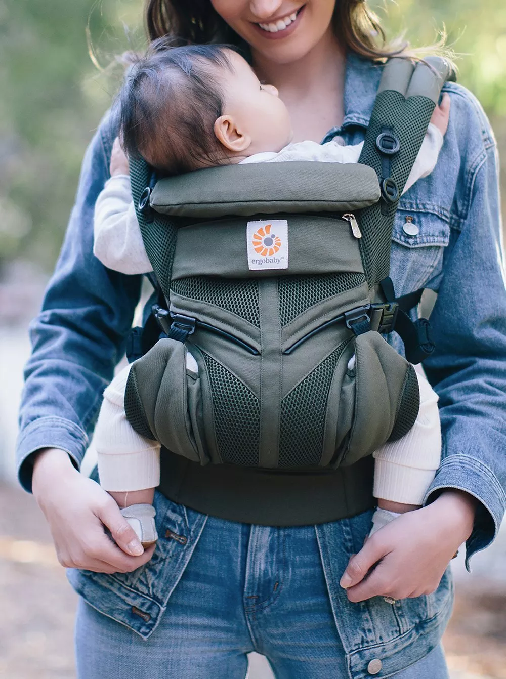 A smiling woman carries a baby in a green ergonomic baby carrier outdoors, wearing a denim jacket.