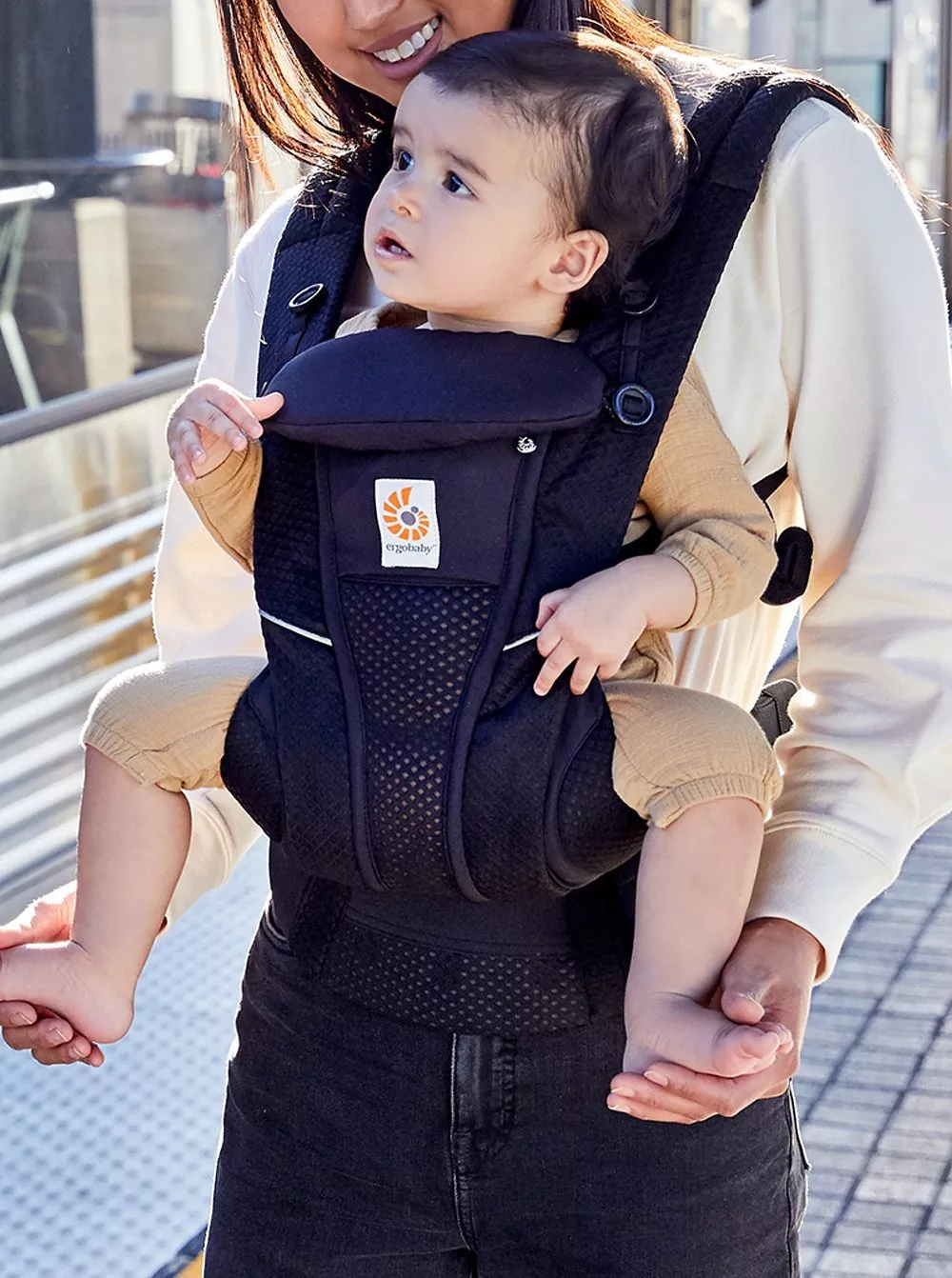 A parent holds a child in a black baby carrier, standing outdoors. The carrier features a distinctive logo.