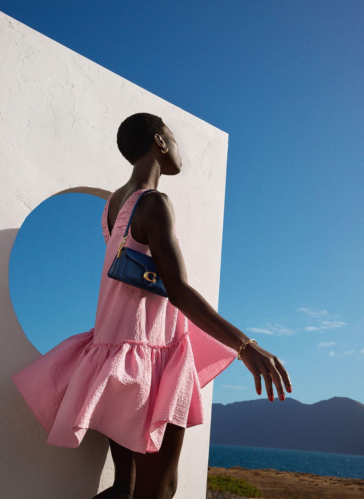 Model wearing pink summer dress, holding a black coach handbag with blue skies and sea view