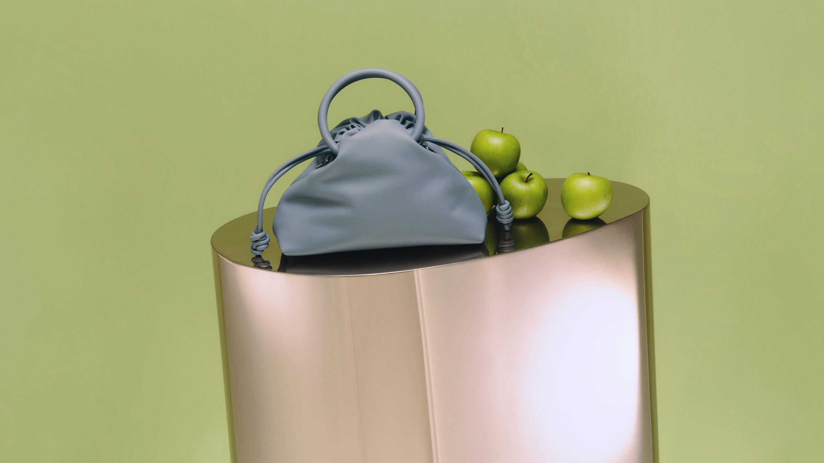 Image of a light blue bag sitting on a stand next to apples with a light green background