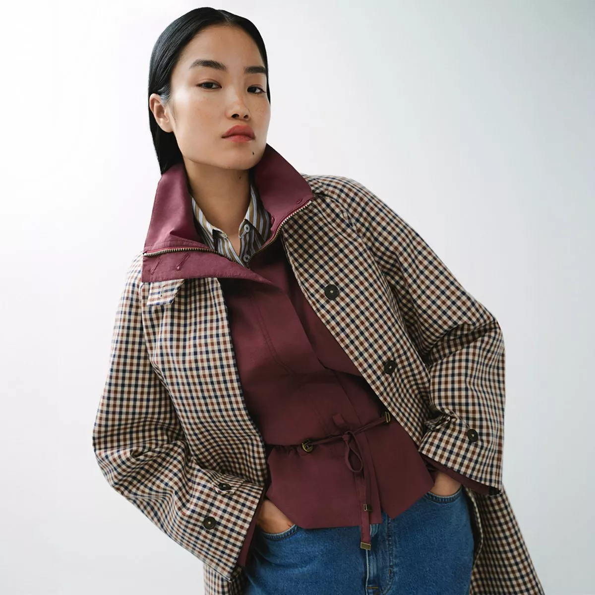woman wearing a plaid trench coat with a high, burgundy-lined collar over a matching burgundy top and blue jeans
