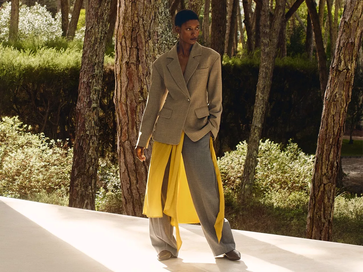 Lady wearing a beige suit with yellow detailing. Standing on a platform in front of trees, in the sun