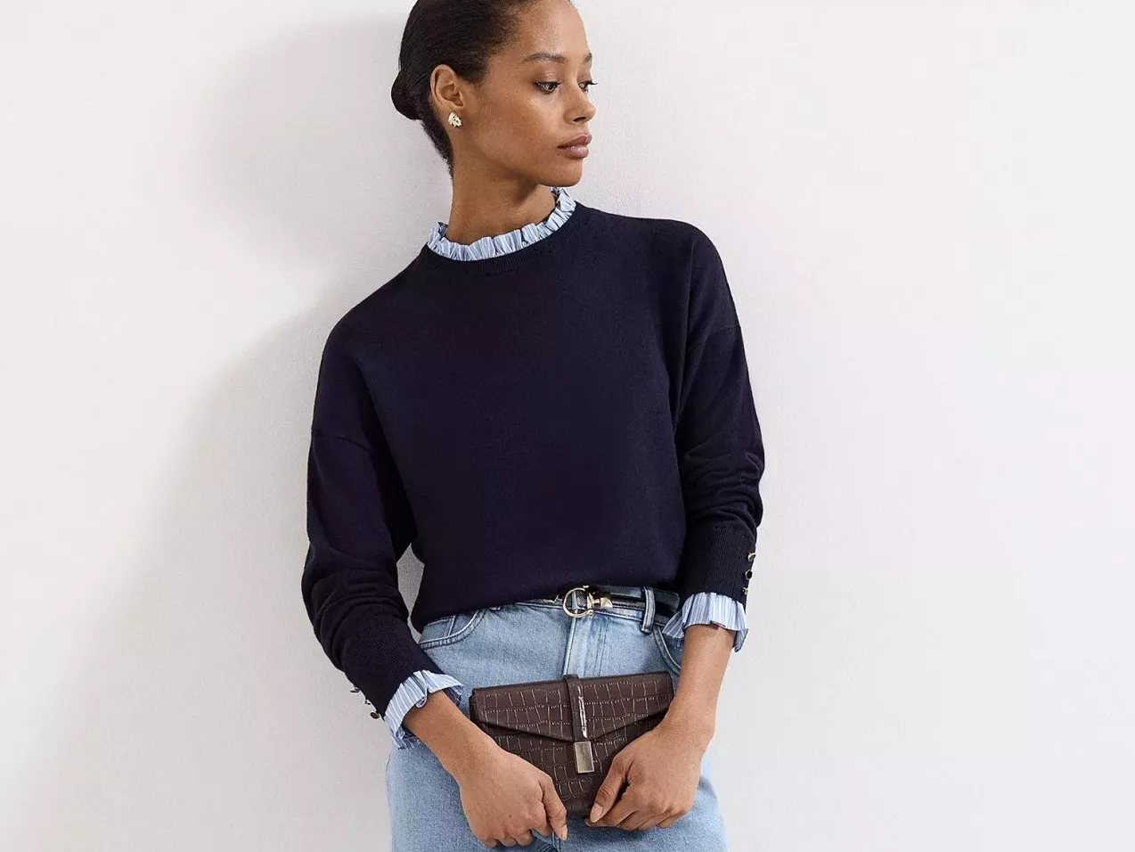 Woman in navy jumper with ruffle collar and light blue jeans, holding brown clutch bag against white wall.
