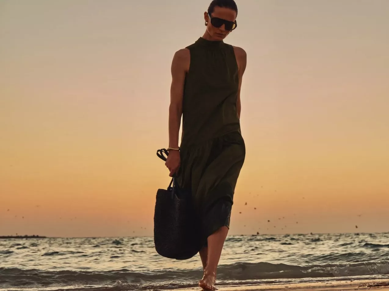 Woman in sunglasses wearing an olive green maxi dress, walking barefoot on a beach at sunset with tote bag
