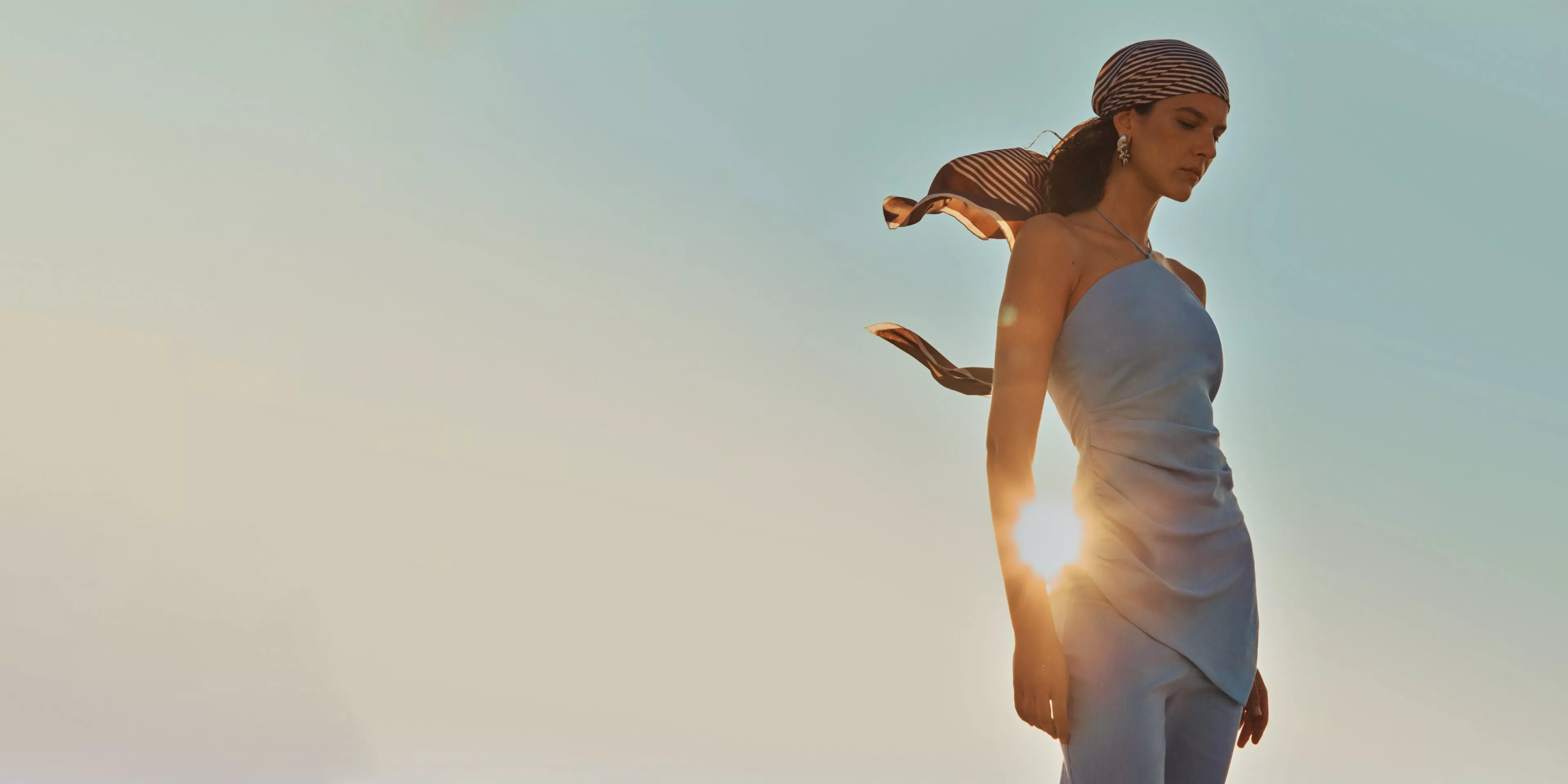 Fashion model in pale blue strapless dress with striped headscarf, backlit by sunset sky outdoors