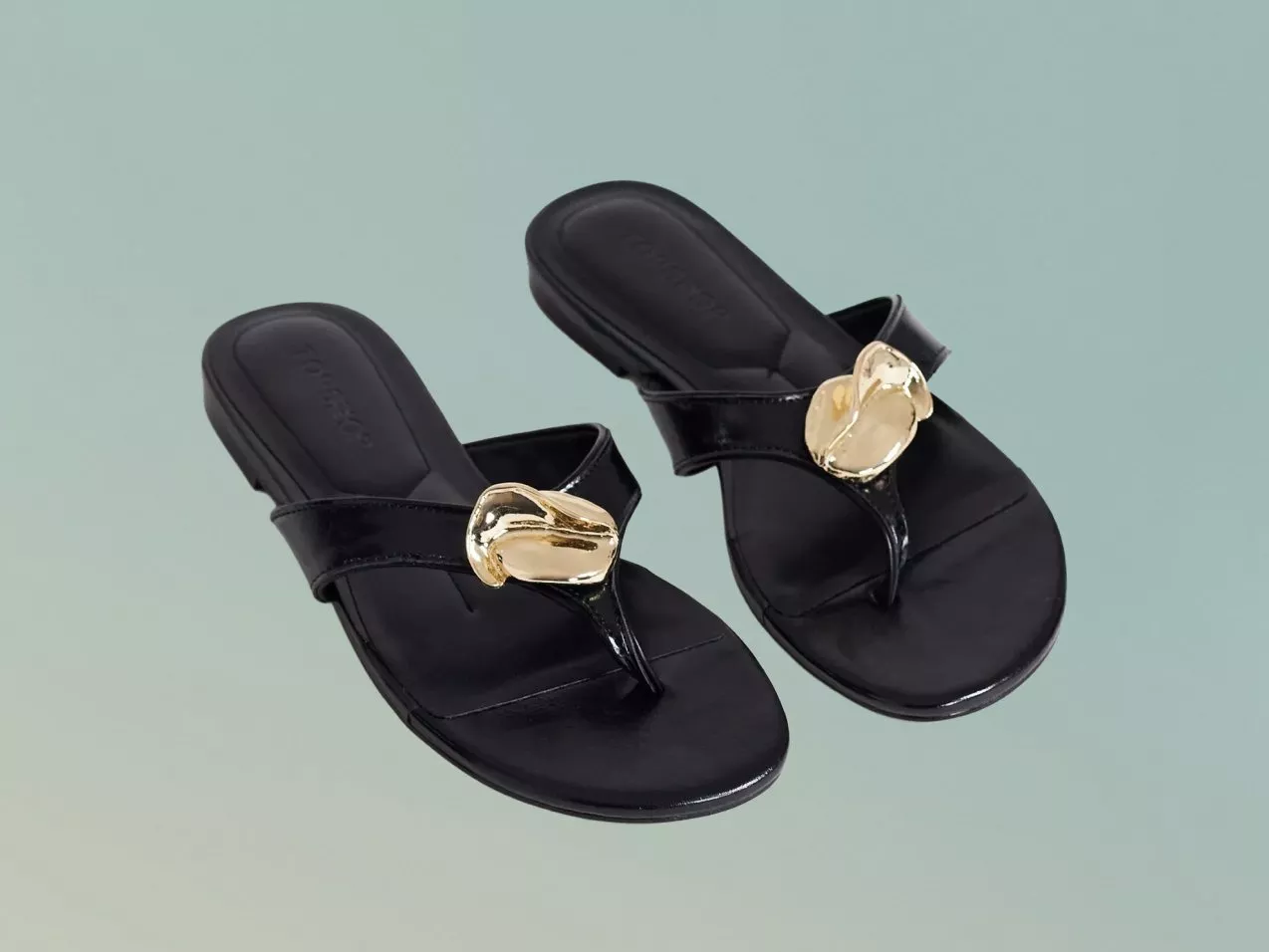 Pair of black women’s slip-on sandals with gold buckle detail, studio product photo on pale green background