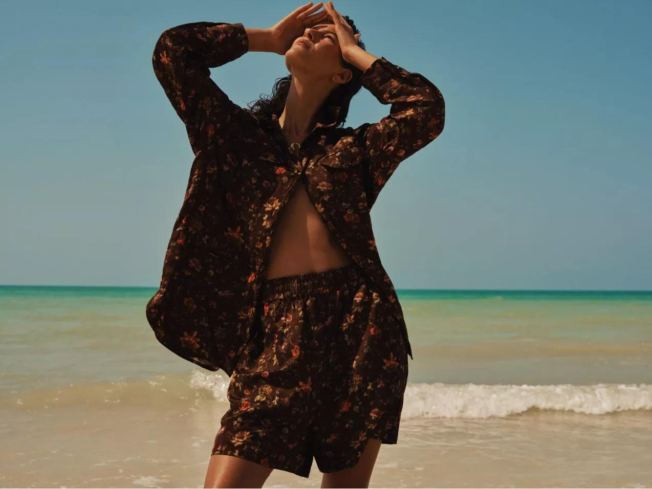 Fashion model posing on a sunny beach in a floral co-ord set, hands raised, sea and blue sky backdrop