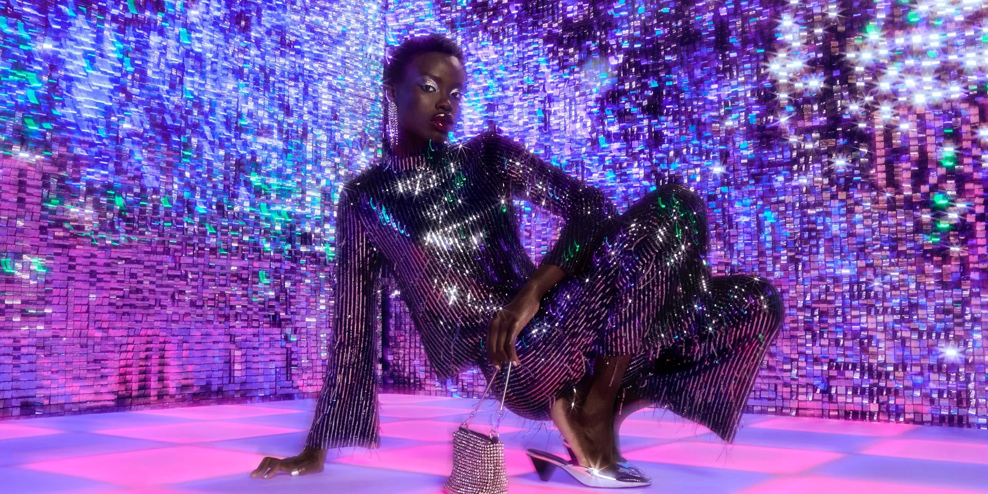 A model wearing a sequin top and matching trousers, crouching down on a pink and purple dancefloor with a purple sequin backdrop