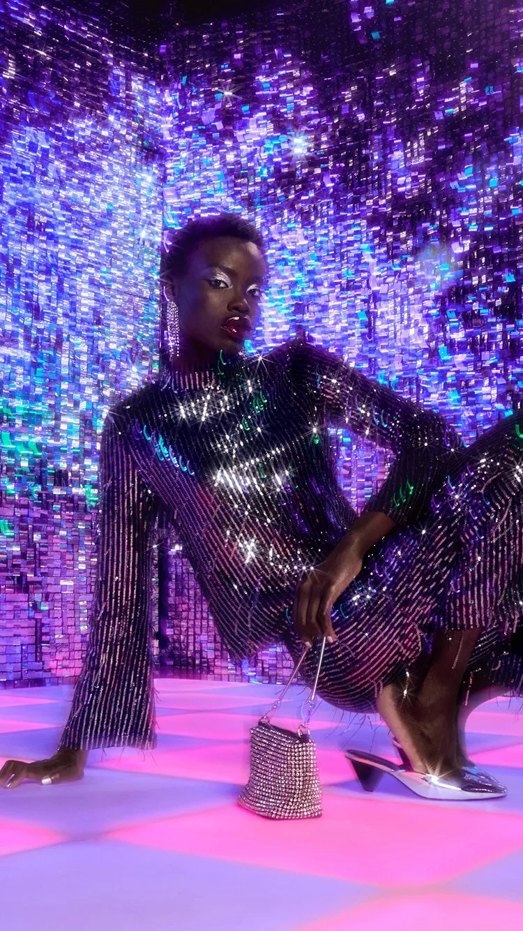 A model wearing a sequin top and matching trousers, crouching down on a pink and purple dancefloor with a purple sequin backdrop