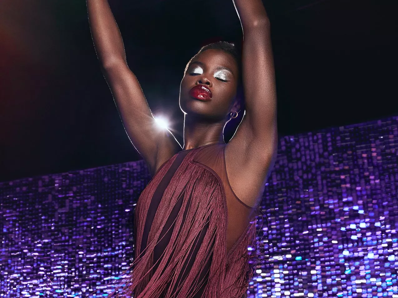 A model wearing a burgandy fringe dress dancing with her arms up wearing red lipstick and silver eye shadow and a purple sequin backdrop