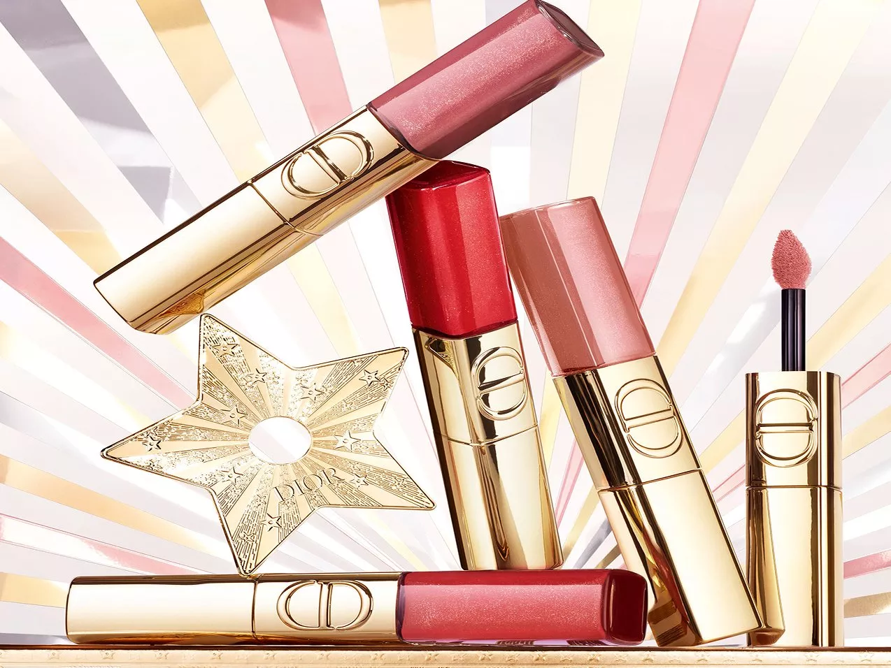 Several Dior lip glosses stacked on each other and a gold star holding them up with a stripy background