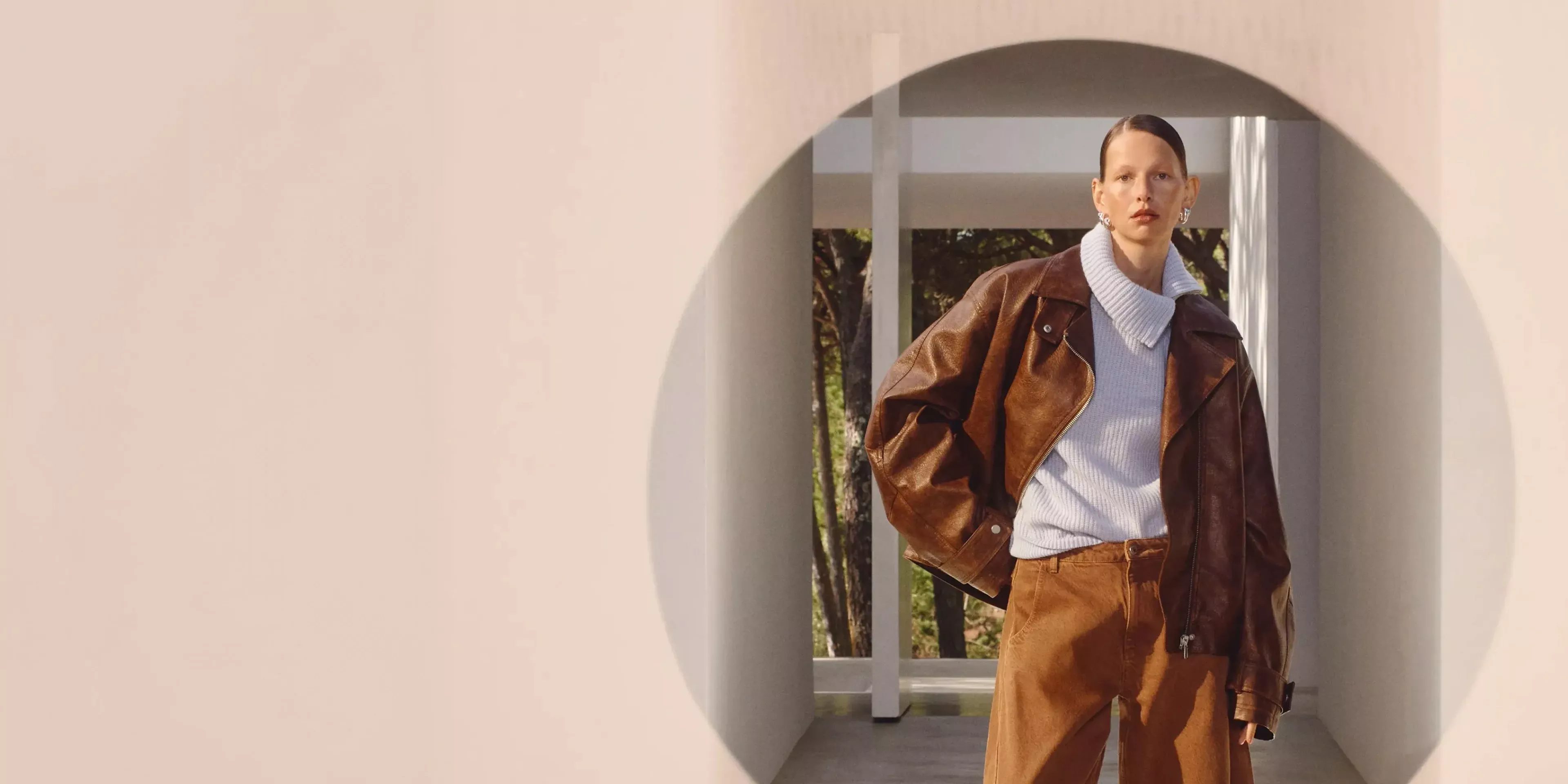 A person in a stylish brown leather jacket and trousers stands in a modern architectural space with natural light.