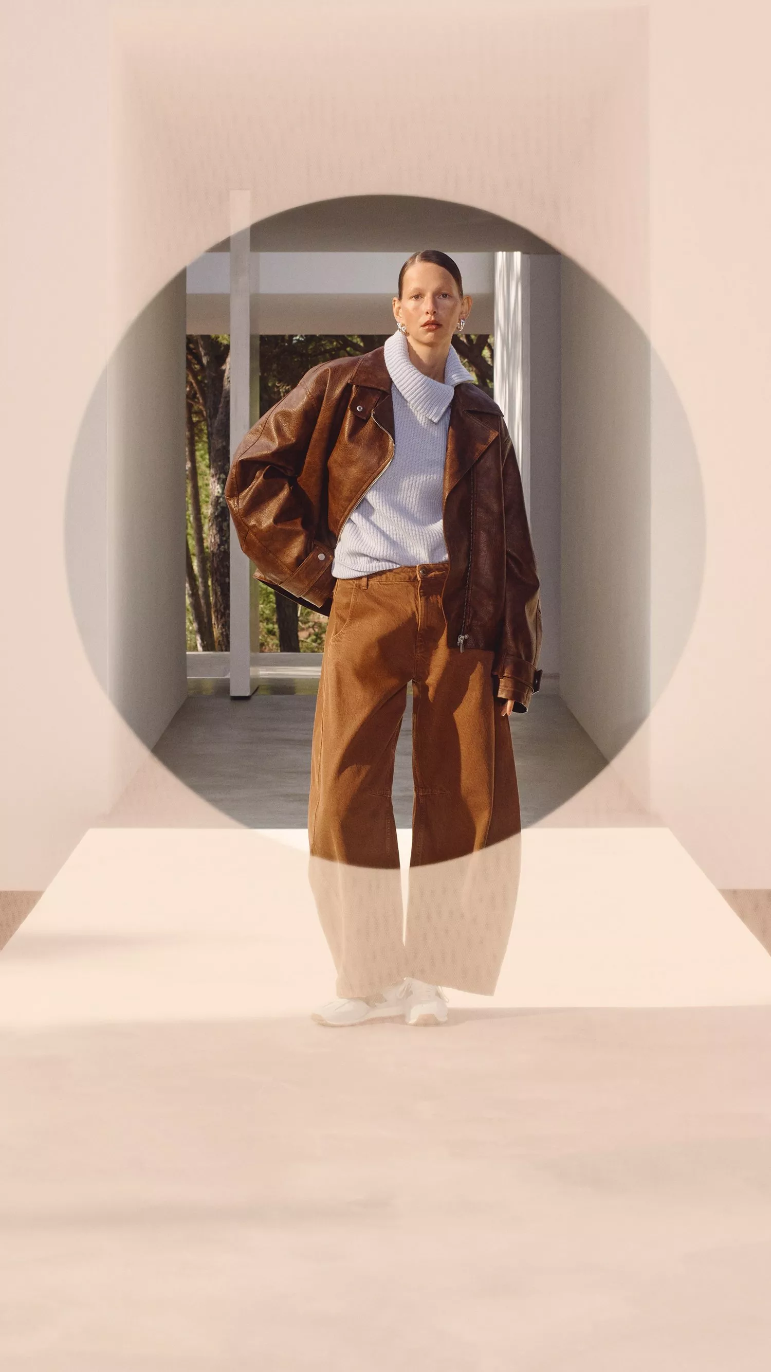 A person wearing a brown leather jacket, white trainers, and wide-leg trousers in a modern, minimalist setting.