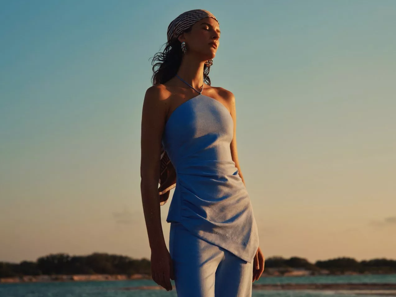 Fashion model in blue halter top and trousers with headscarf, posing by the sea at golden hour sunset