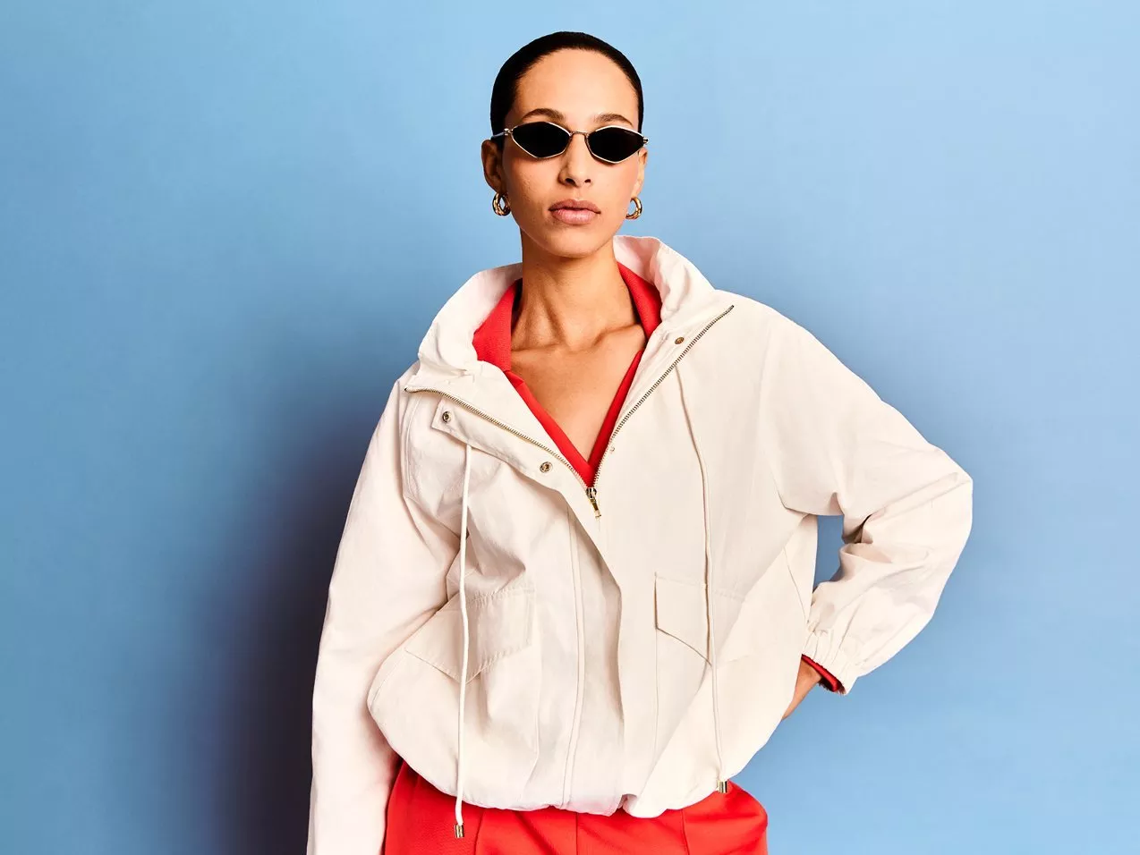 Woman in white hooded jacket, red trousers and black sunglasses,  against a blue background