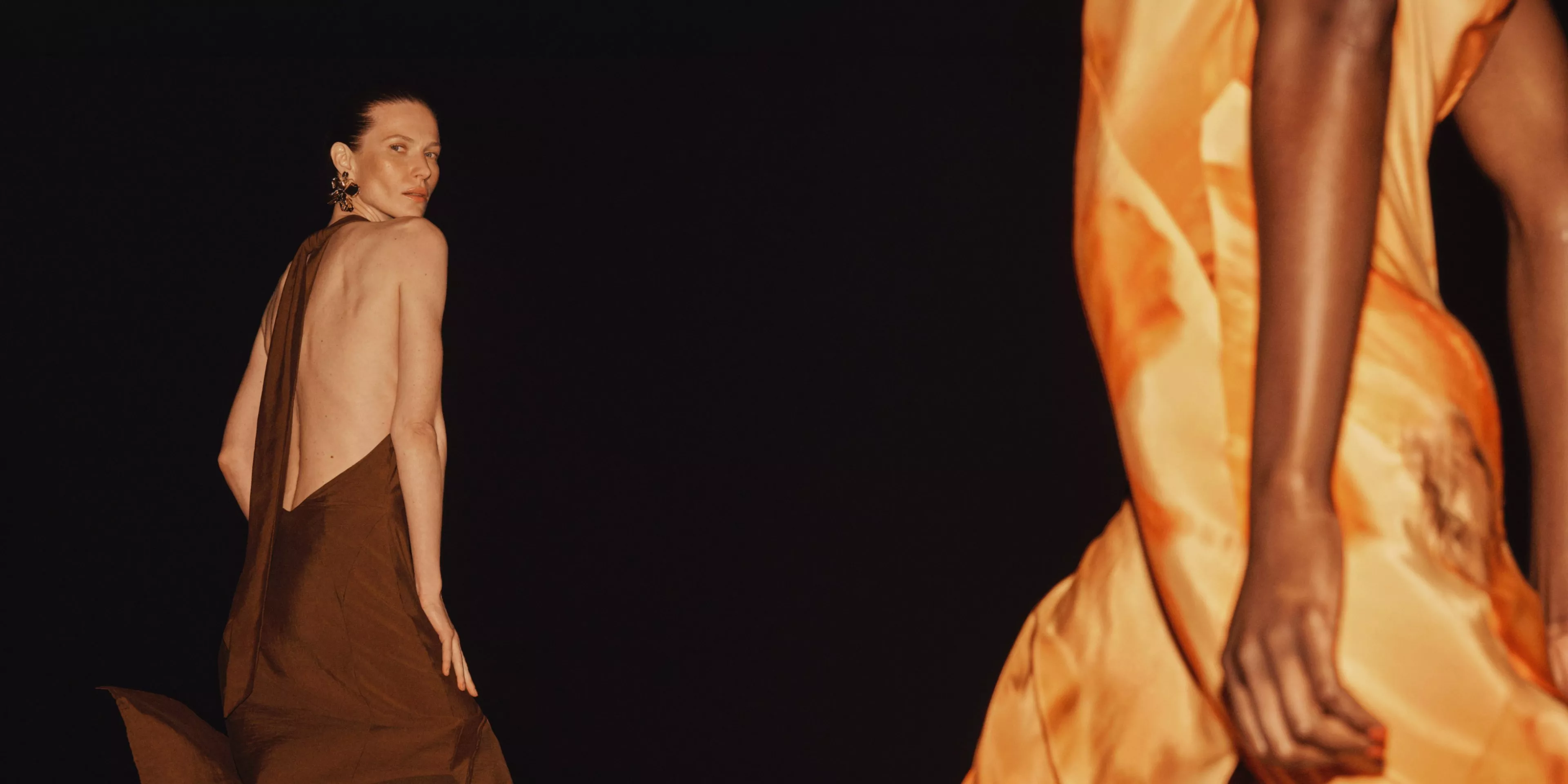 Models in elegant evening summer dresses, one backless satin brown dress and one orange satin dress