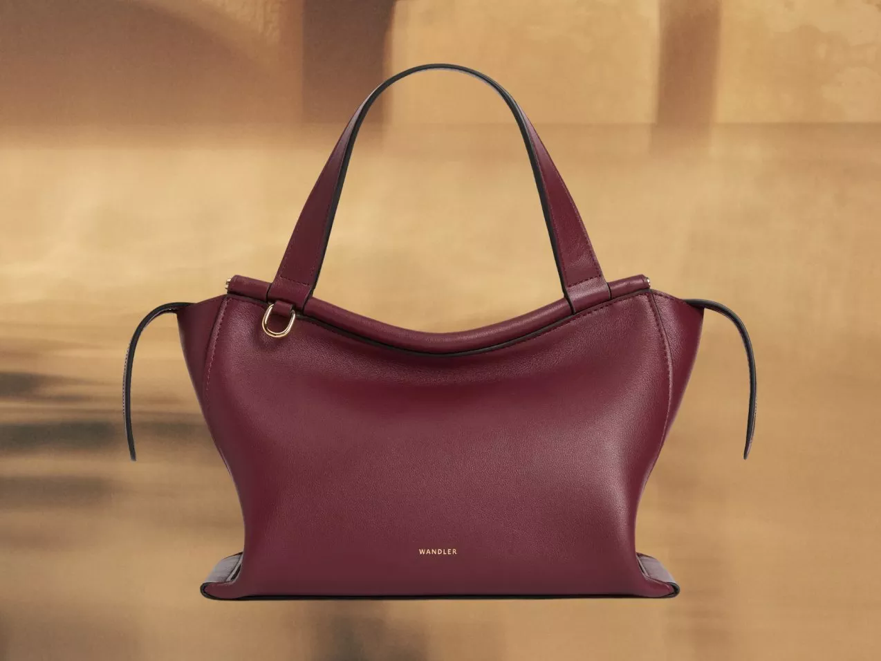 Burgundy leather Wandler tote handbag with top handles and gold ring detail, on beige background