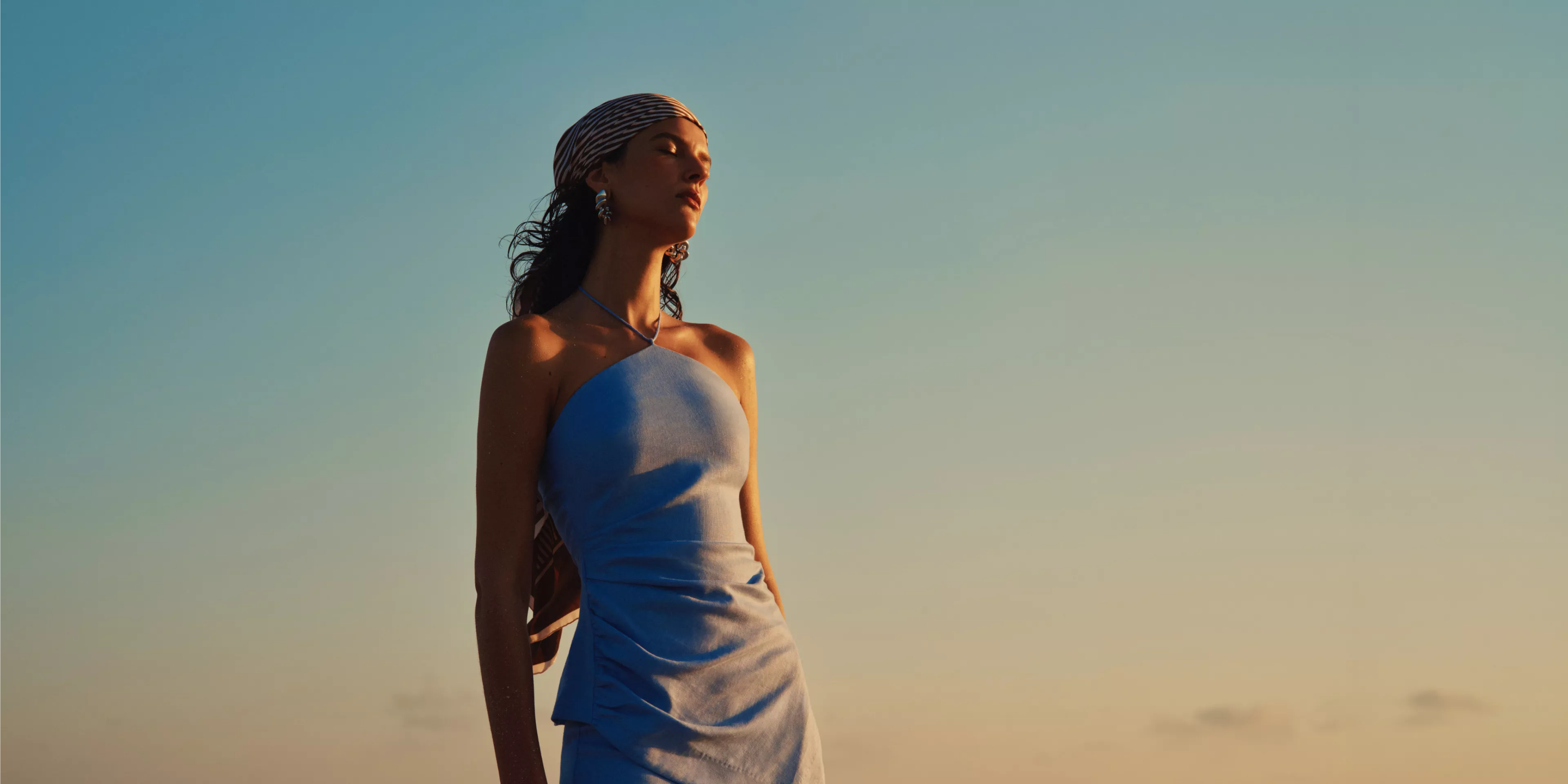 Fashion portrait of woman in light blue dress and headscarf, standing in warm sunset light against clear sky