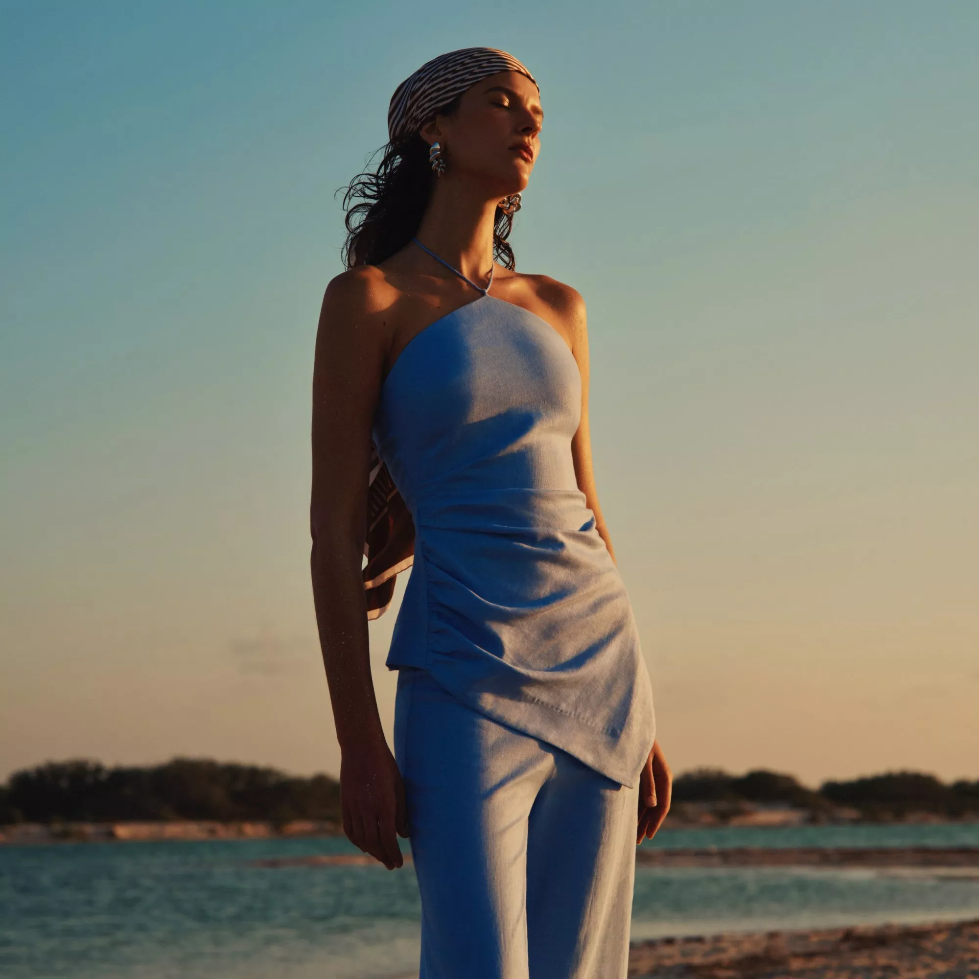 Fashion model in light blue halter top and trousers with headscarf, posing by the sea at sunset