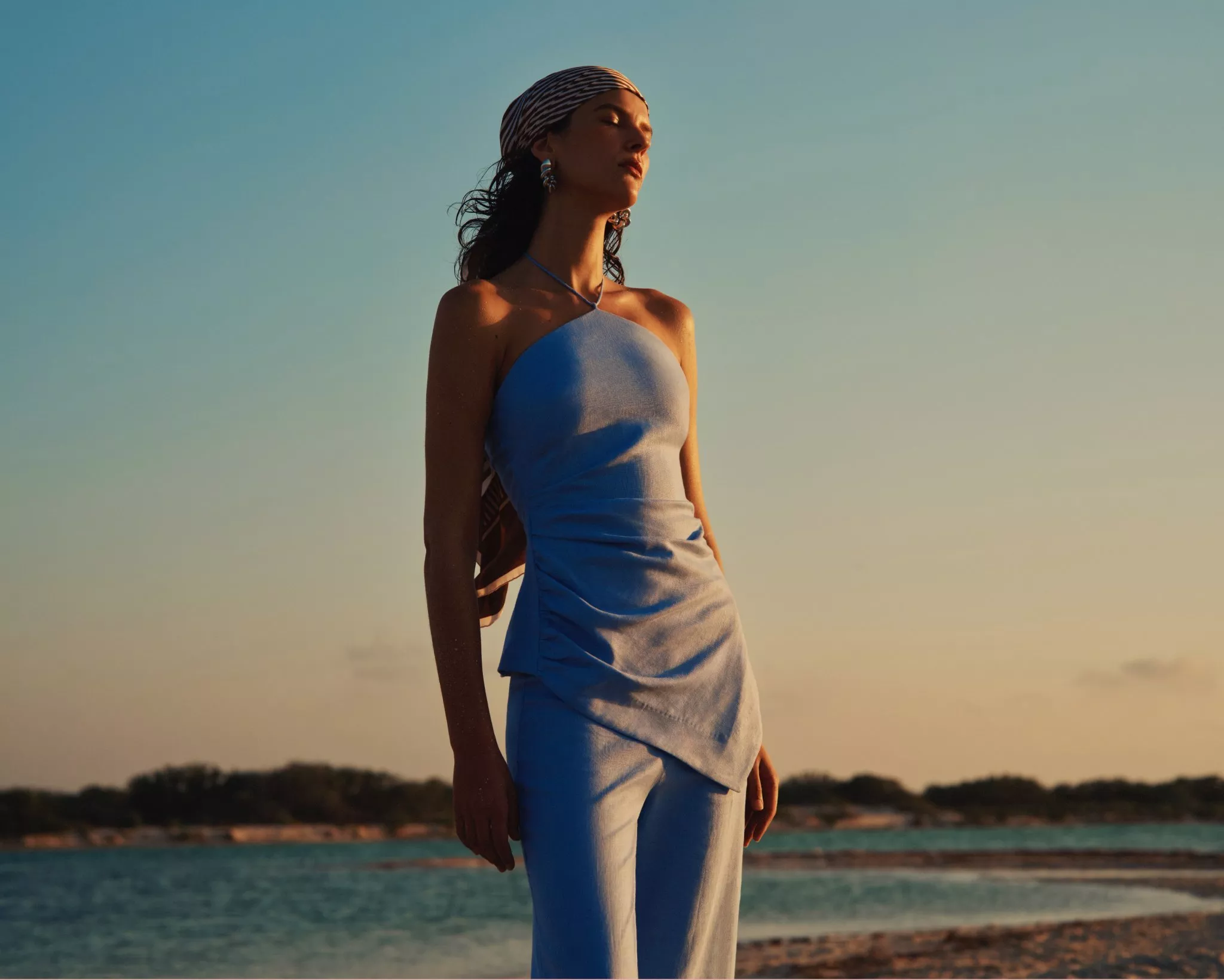 Woman wearing head scarf and strapless top, against a sea backdrop 