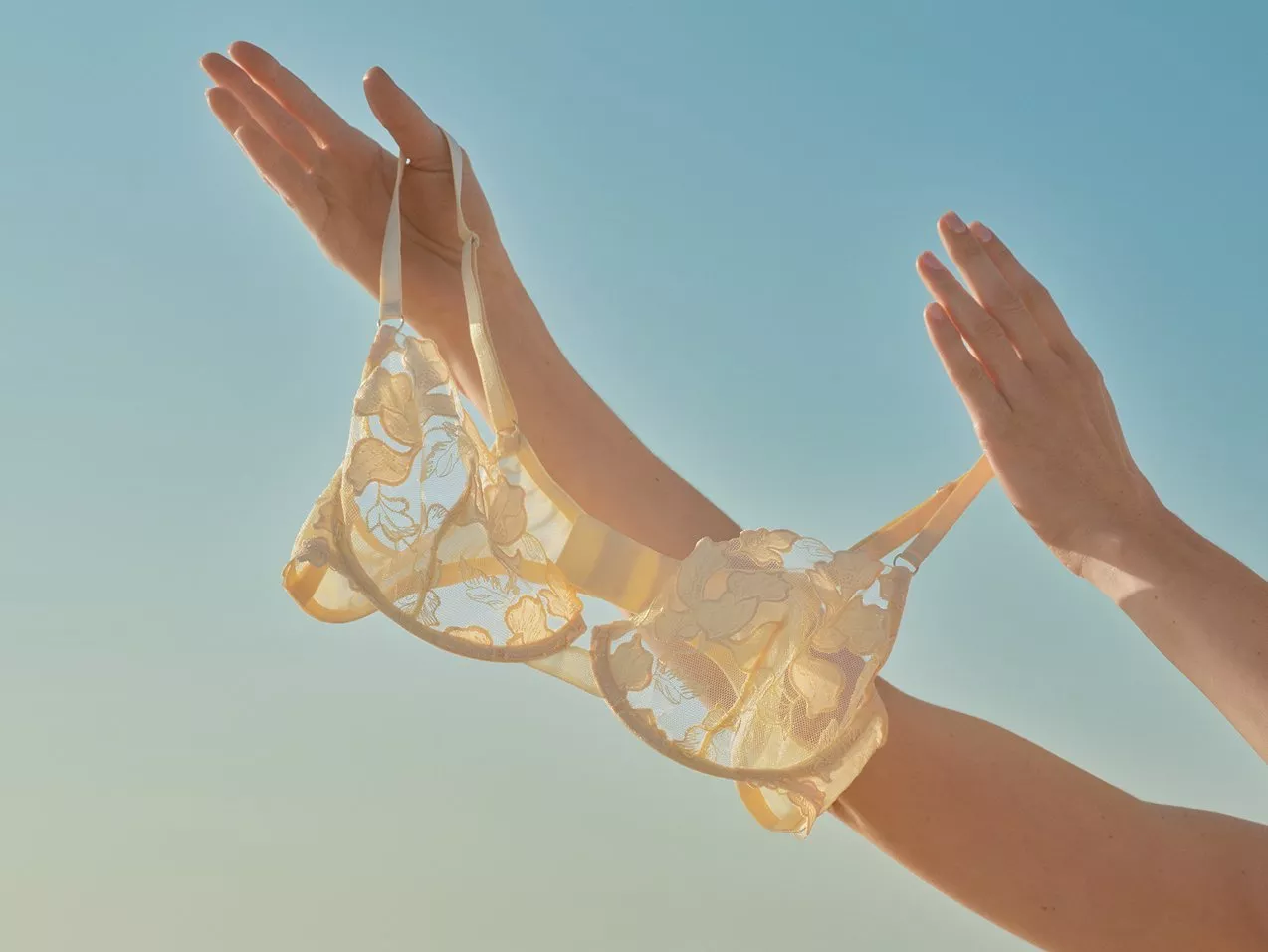 Hands holding a cream coloured lace bra against a clear blue sky