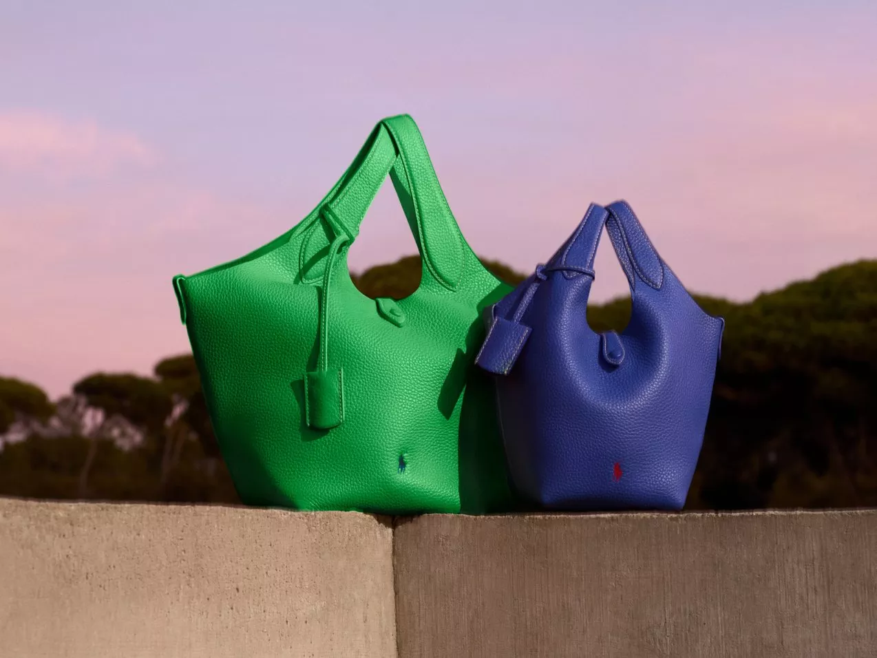 Two stylish leather handbags, one green and one blue, are placed on a concrete surface against a serene sunset sky.