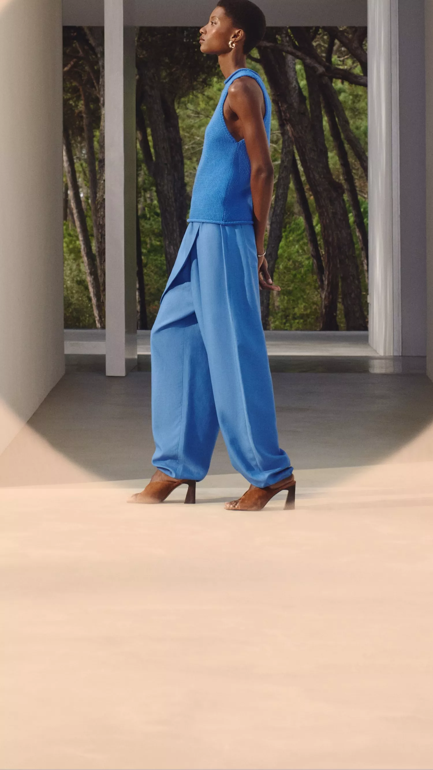Fashion model in vibrant blue sleeveless top and flowing trousers, walking confidently in a modern setting.