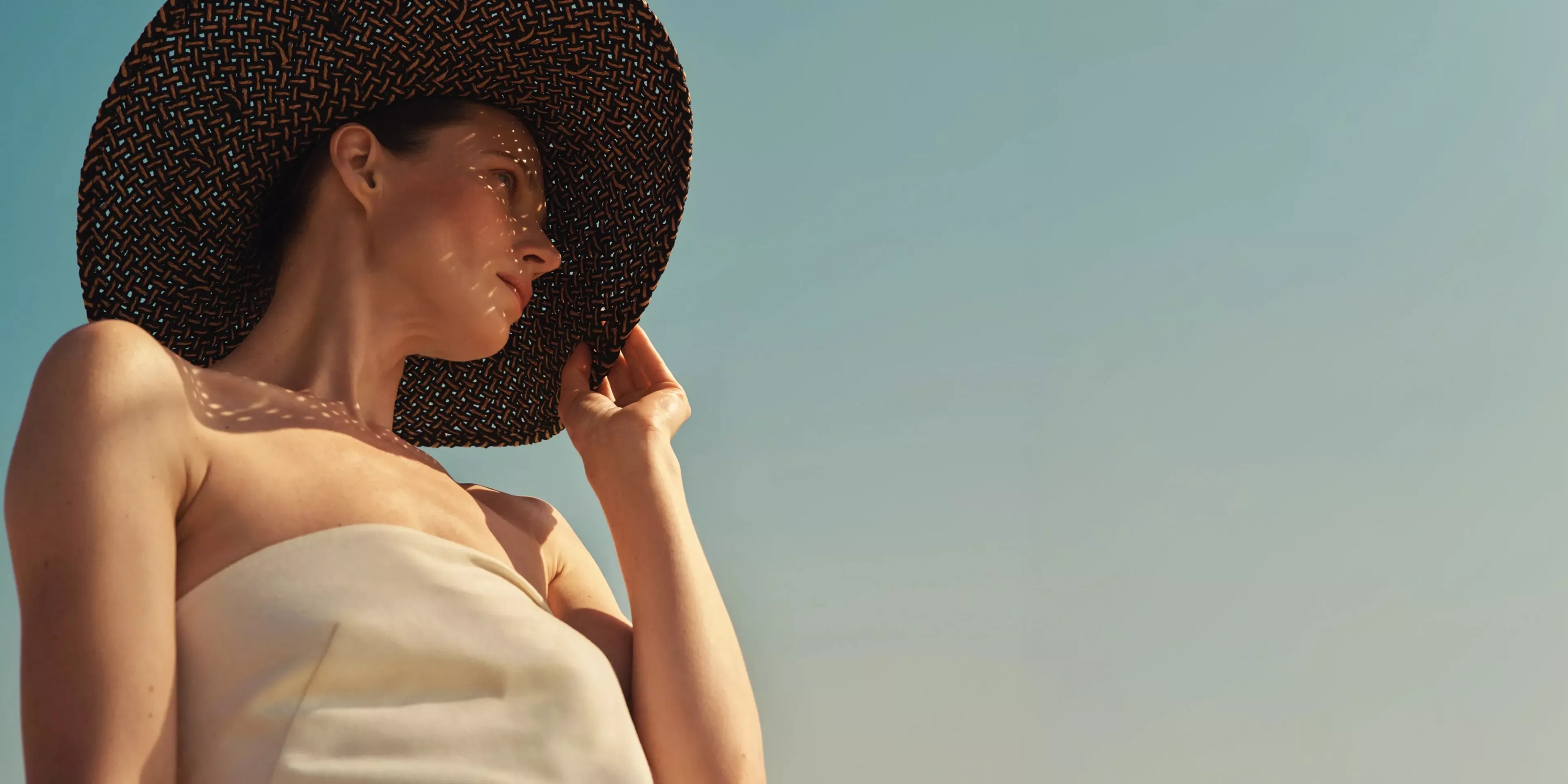 Woman in strapless summer dress wearing a wide-brim straw hat, sunlit portrait against clear sky