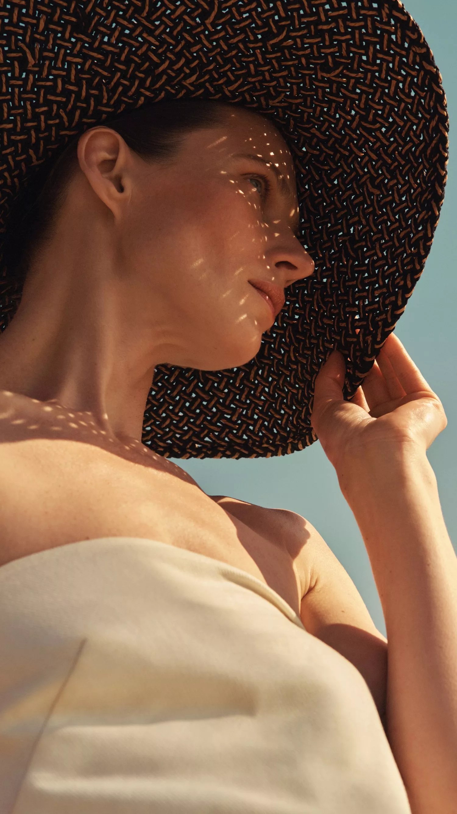 Woman in strapless dress wearing wide-brim woven sun hat, enjoying summer sunshine against clear blue sky