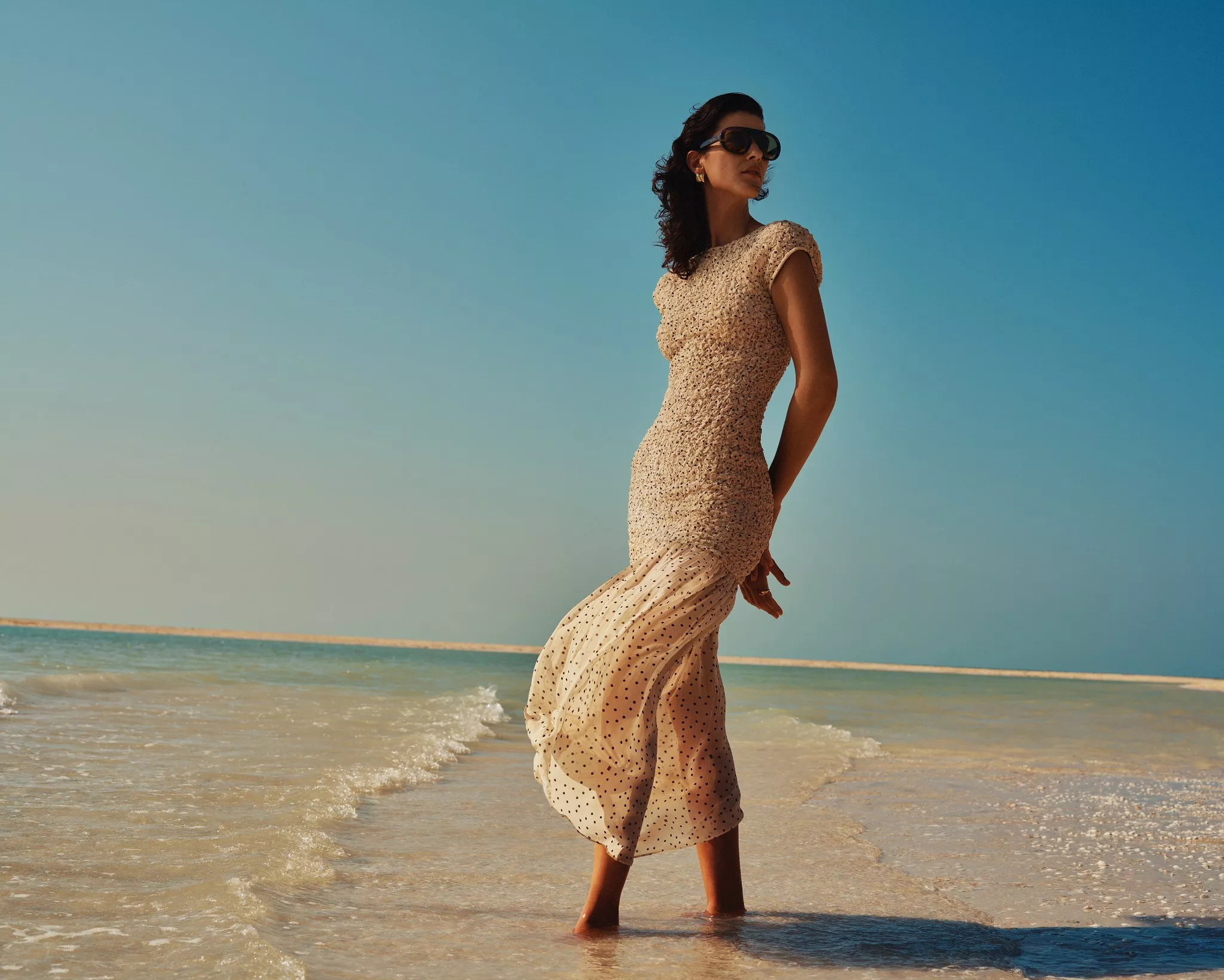 Woman in sunglasses wearing a beige summer dress standing in shallow sea water on a sunny beach shoreline