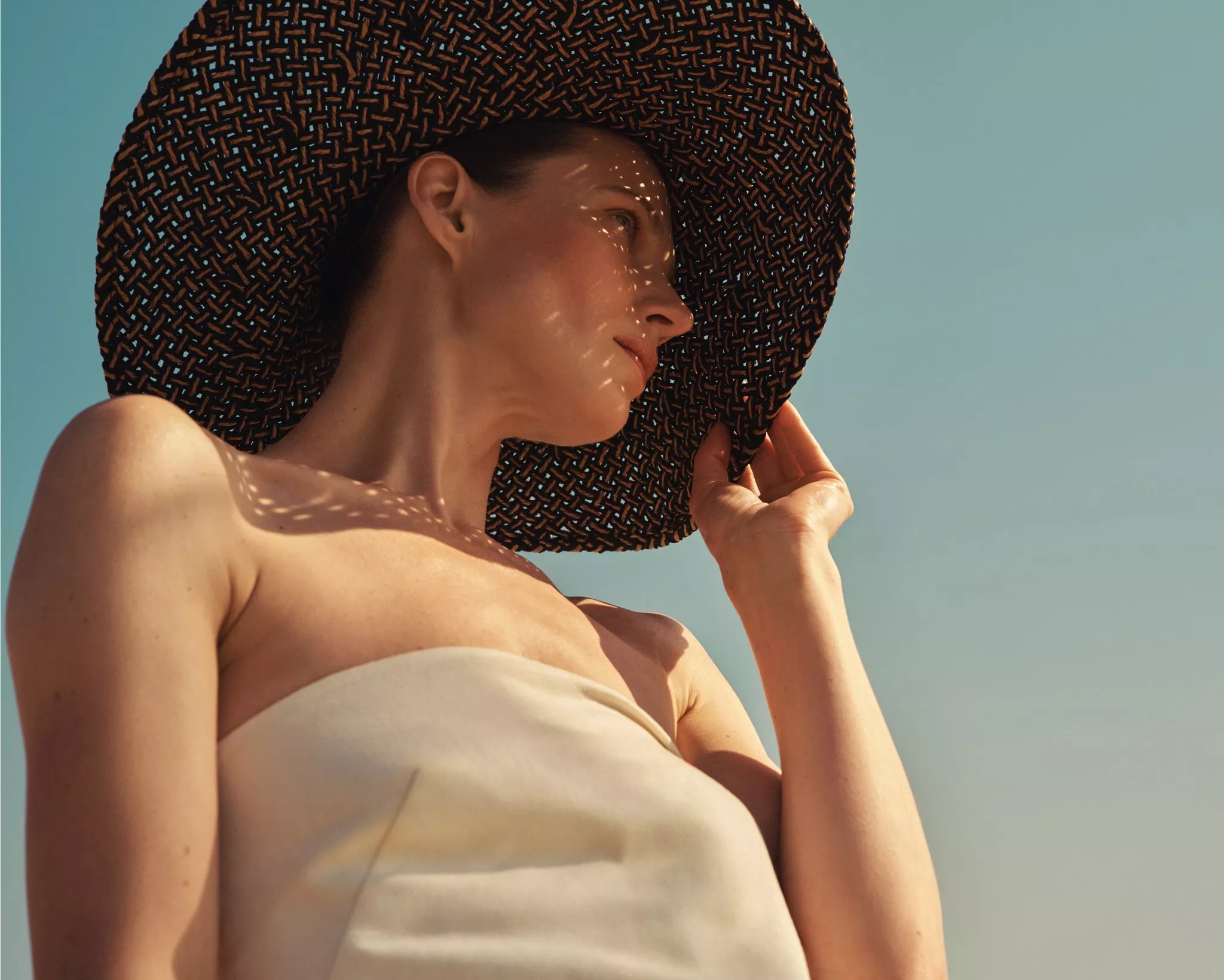 Woman in strapless summer dress wearing a wide-brim woven sun hat, sunlit portrait against clear blue sky