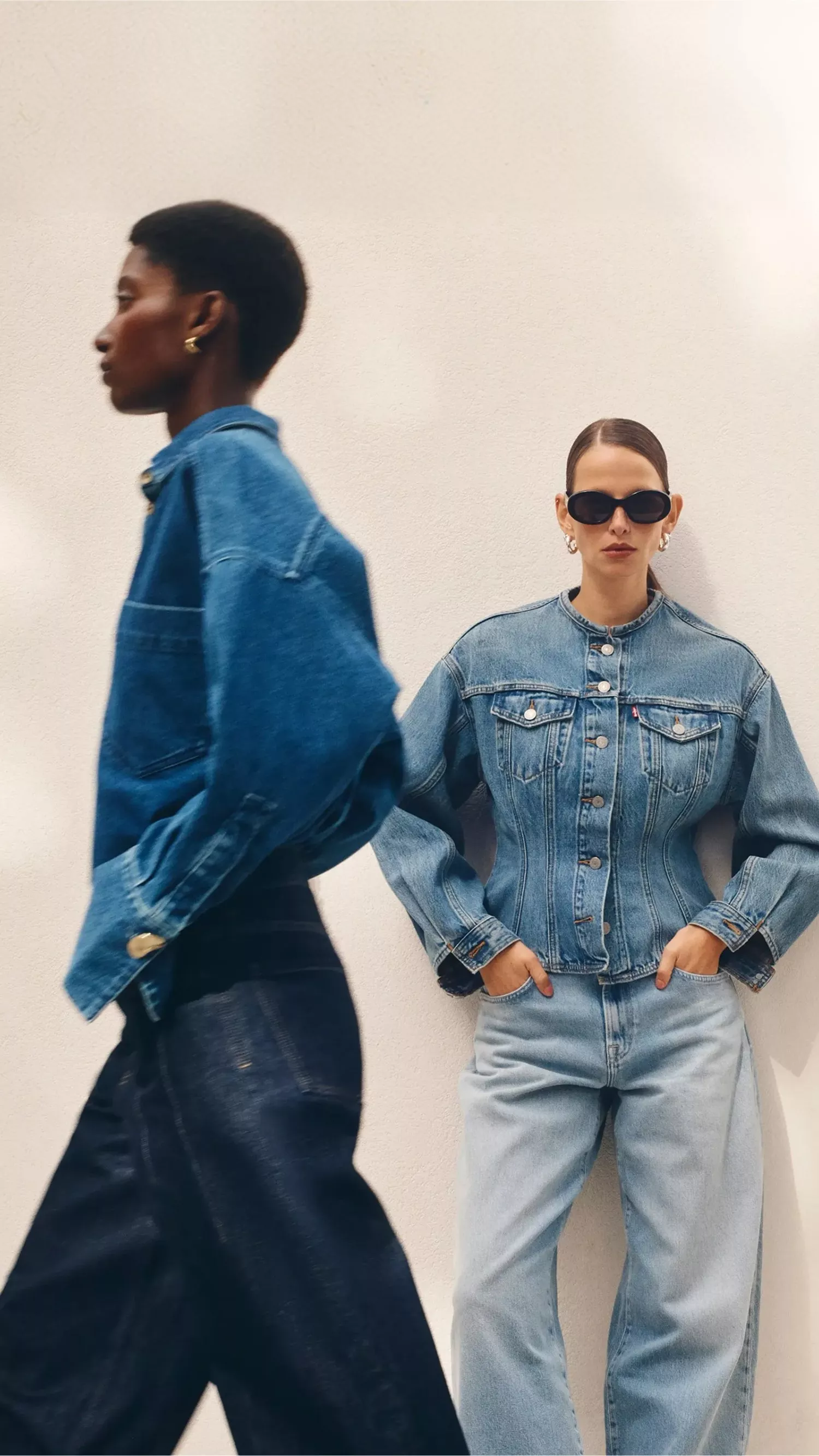 Two women pose in denim; one is in profile wearing a dark denim shirt-jacket, while the other faces forward wearing a tailored light-wash denim jacket and dark sunglasses.