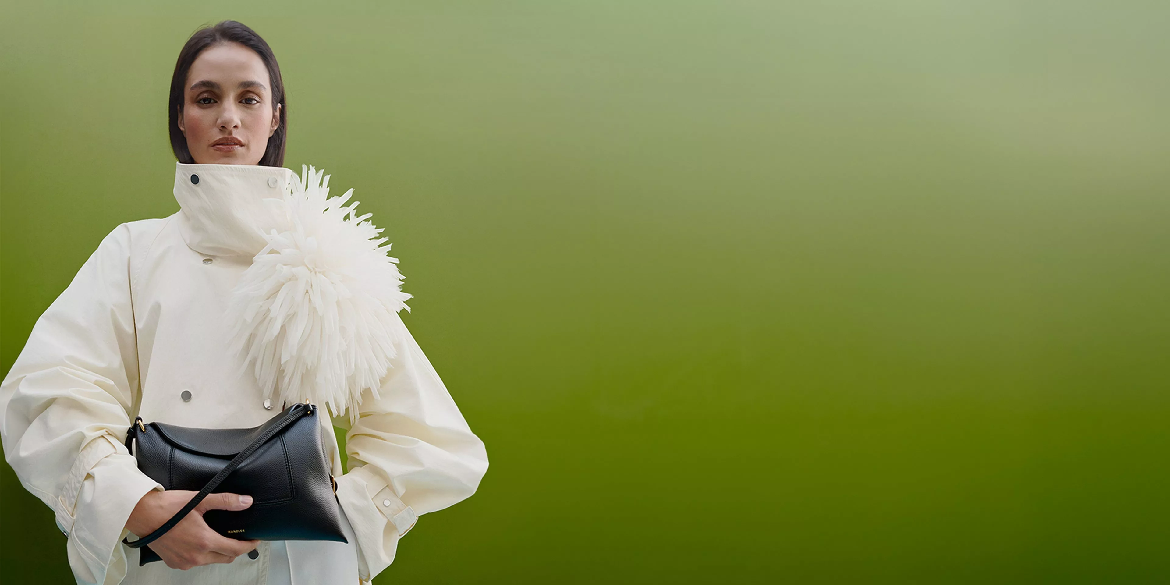 Fashion model in white high-collar coat with feather detail, holding black clutch bag against green backdrop