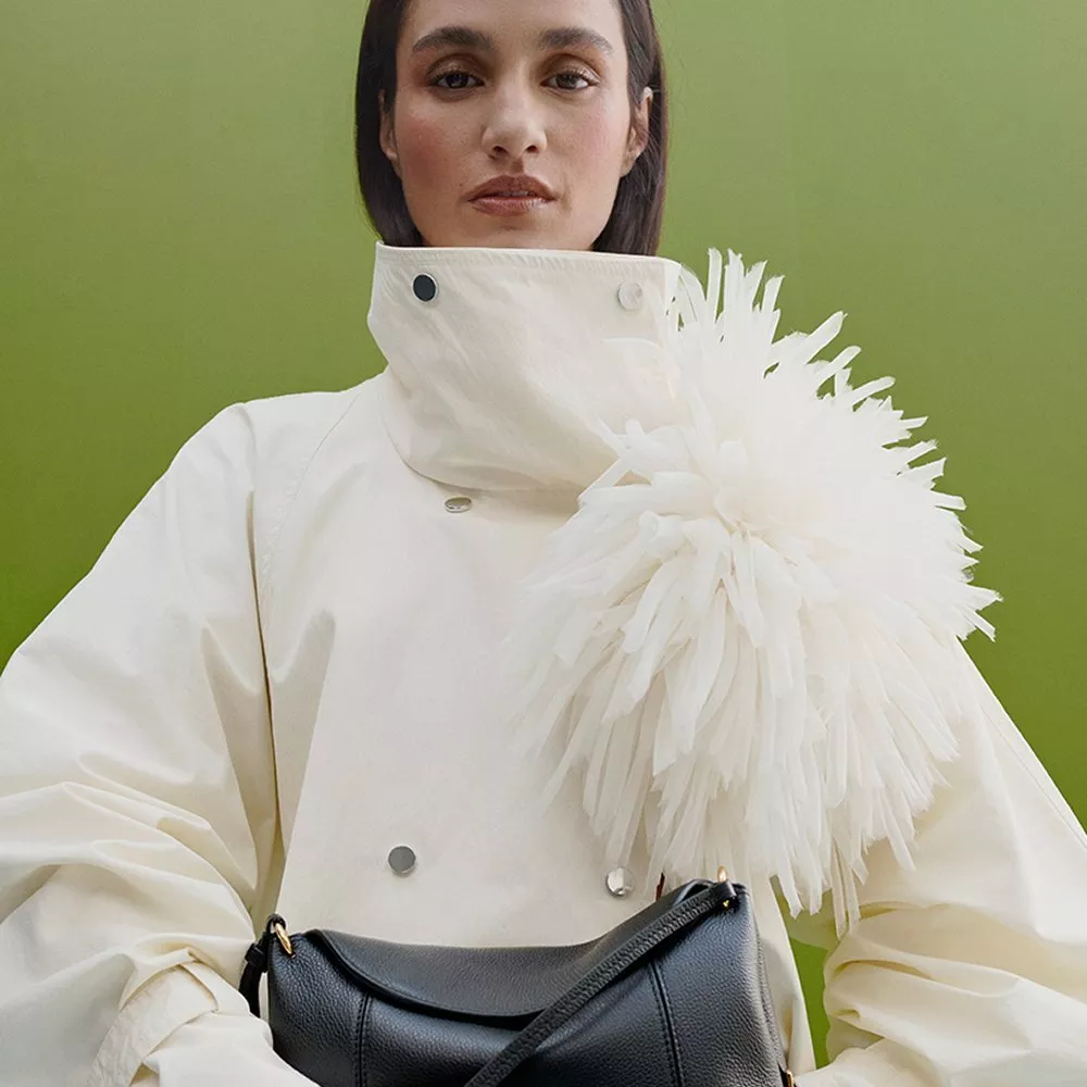 Fashion portrait of woman in white high-collar coat with feather detail, holding black leather handbag on green backdrop