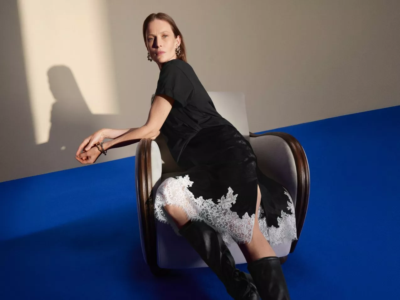 Fashion portrait of a woman in black dress with white lace hem, seated in armchair against blue backdrop.